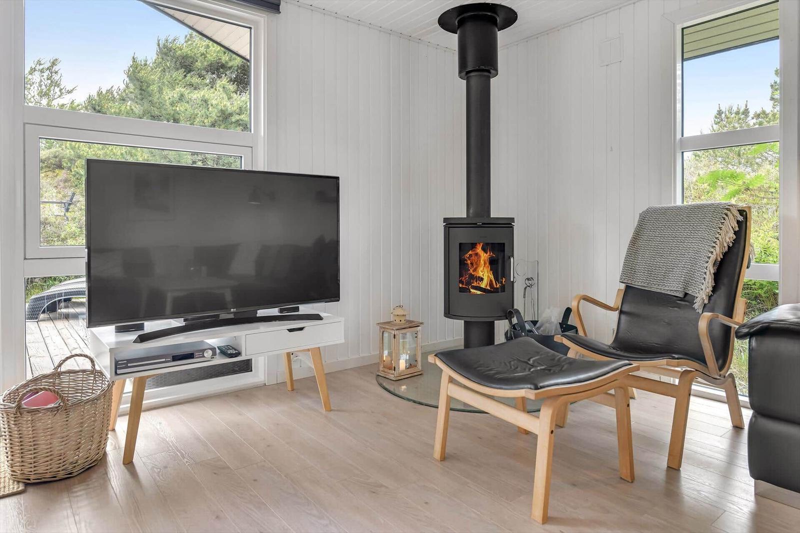 Wohnzimmer mit Kamin, Fernseher und Sitzgelegenheiten. Blick auf grüne Umgebung.