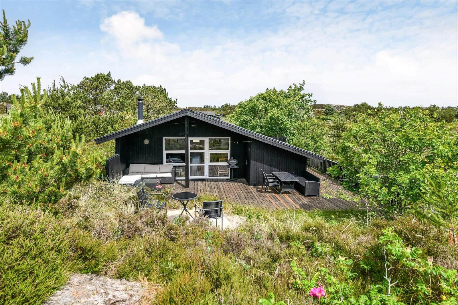 Schwarzes Ferienhaus mit Holzterrasse und Gartenmöbeln in grüner Umgebung.