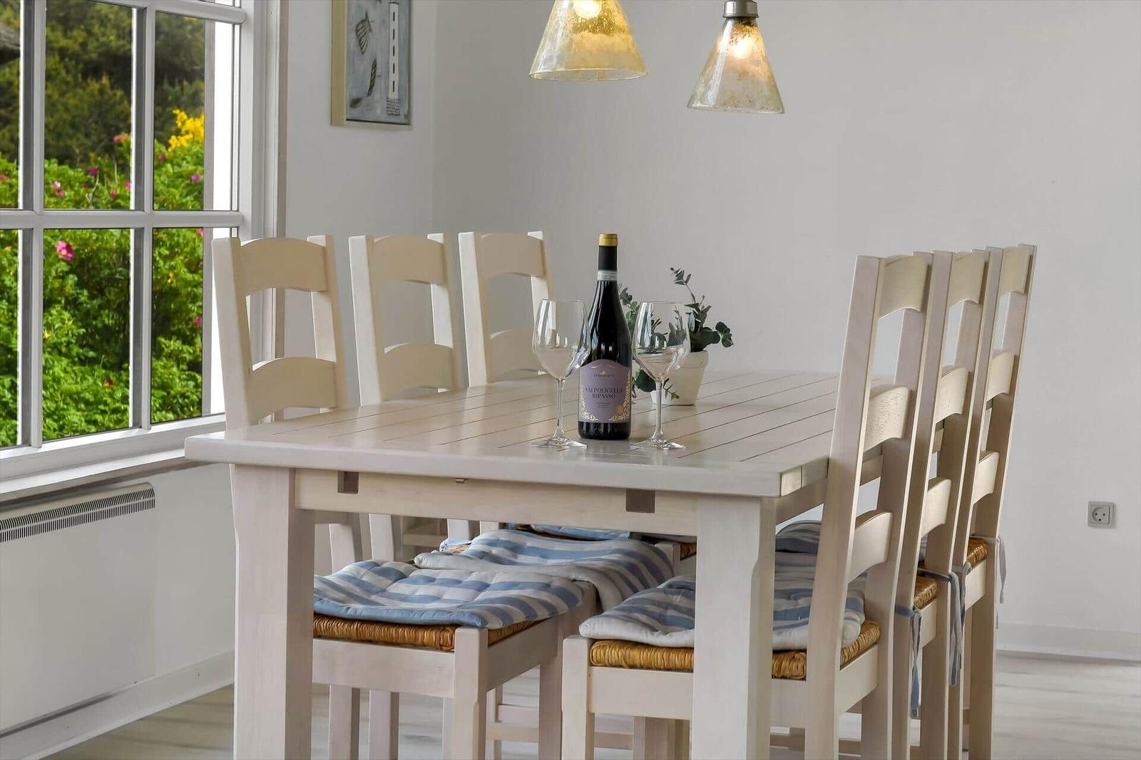 Dining table with six chairs and wine bottle by window with garden view.