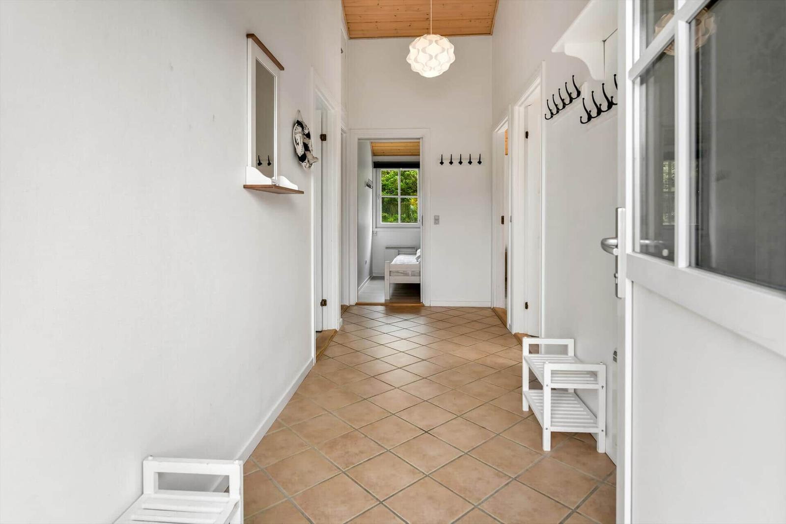 Hallway with terracotta floor, white walls, and hooks. Bedroom with window visible in the back.