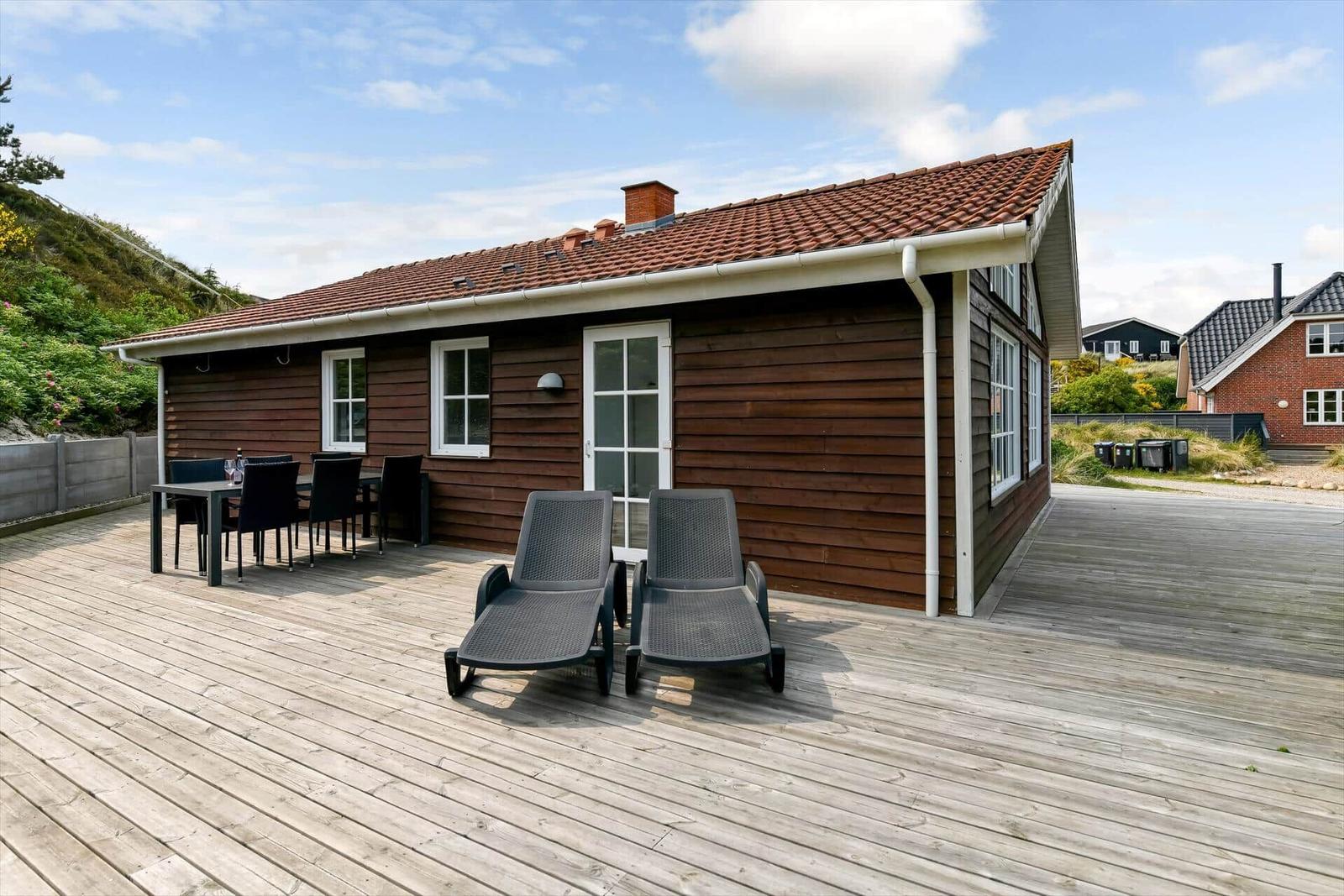 Wooden house with terrace, loungers, and table. Background: greenery and neighboring houses.
