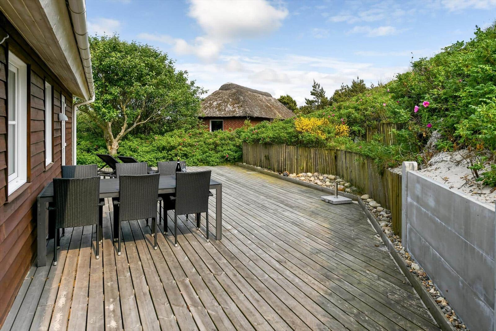Wooden terrace with table and chairs, view of greenery and a thatched roof in the background.