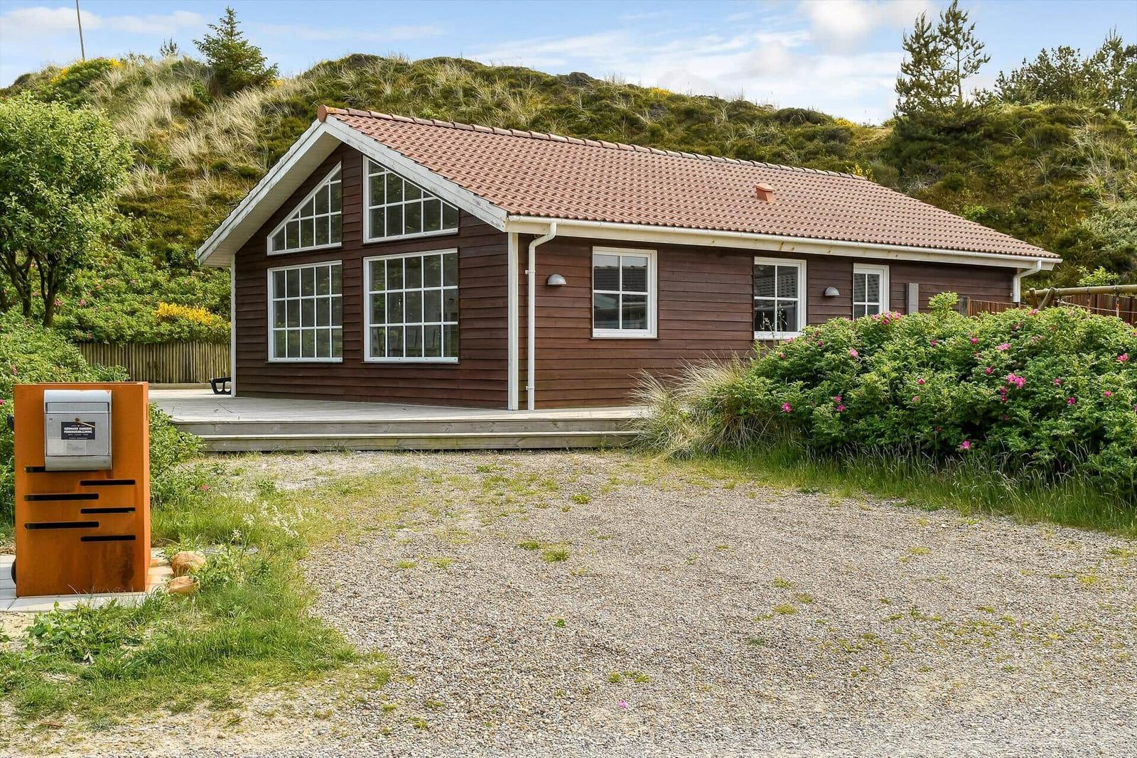 A brown wooden house with a red roof and white window frames stands on a gravel plot.