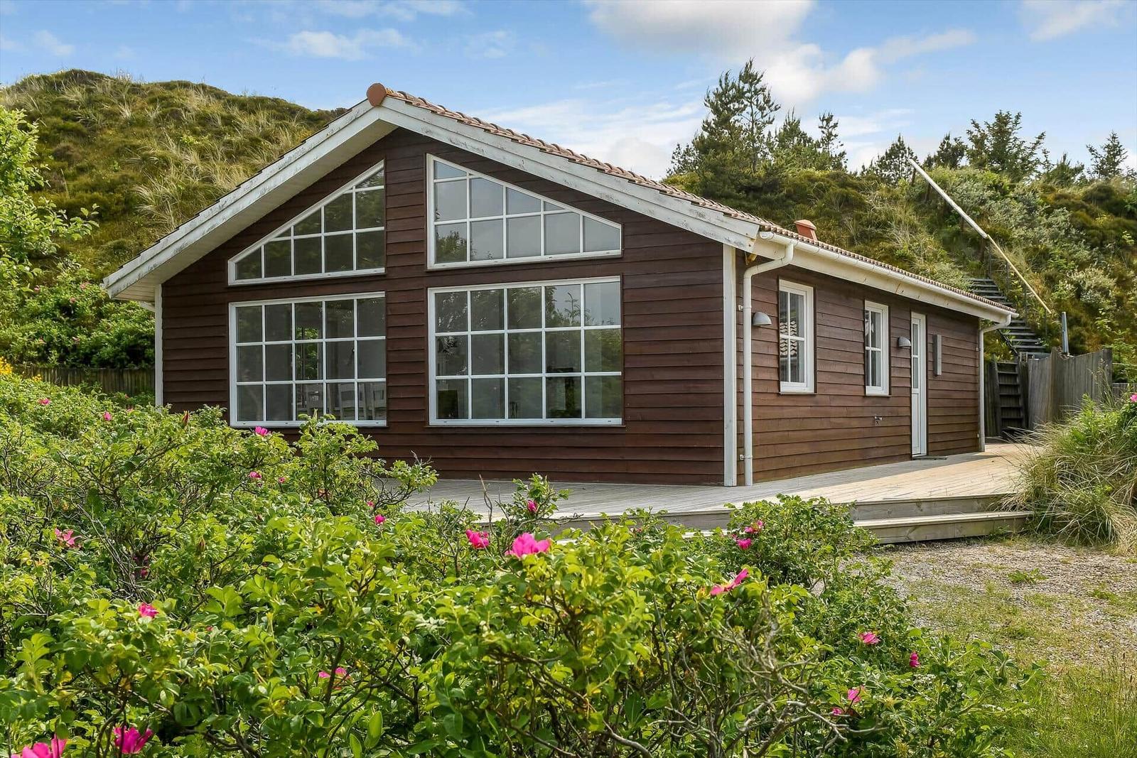 Wooden house with large windows and terrace in a green setting.
