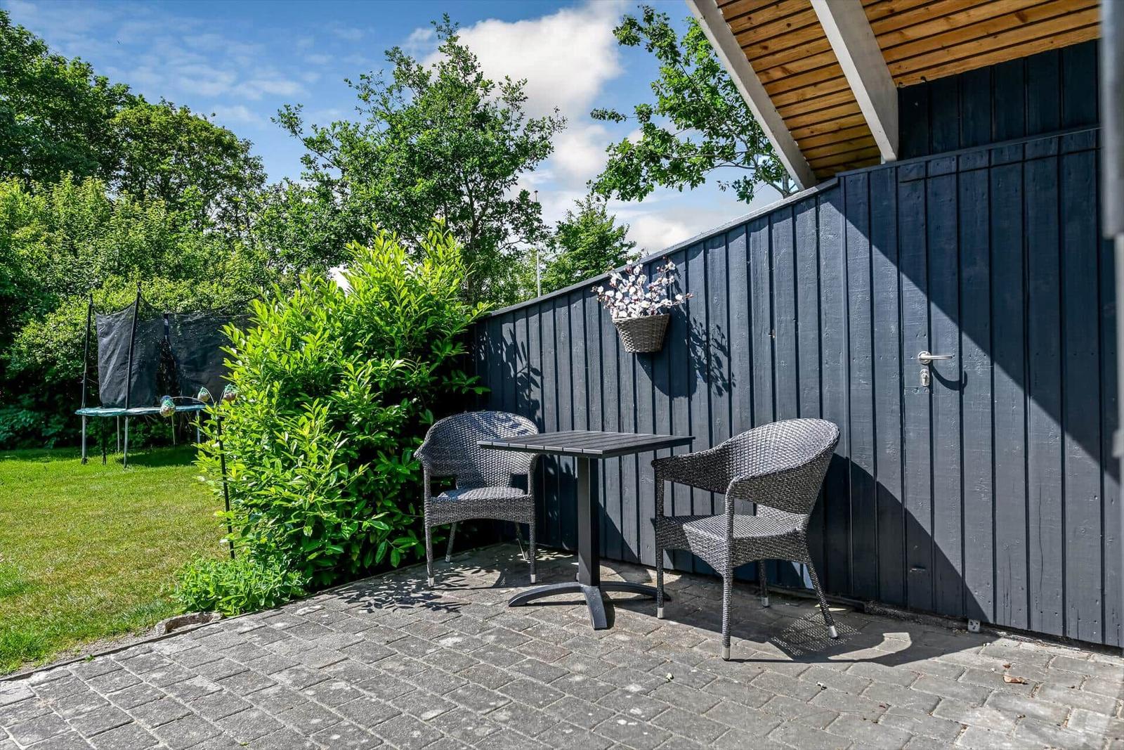 Terrasse mit Tisch und zwei Stühlen, umgeben von Grün und einem dunklen Holzzaun.