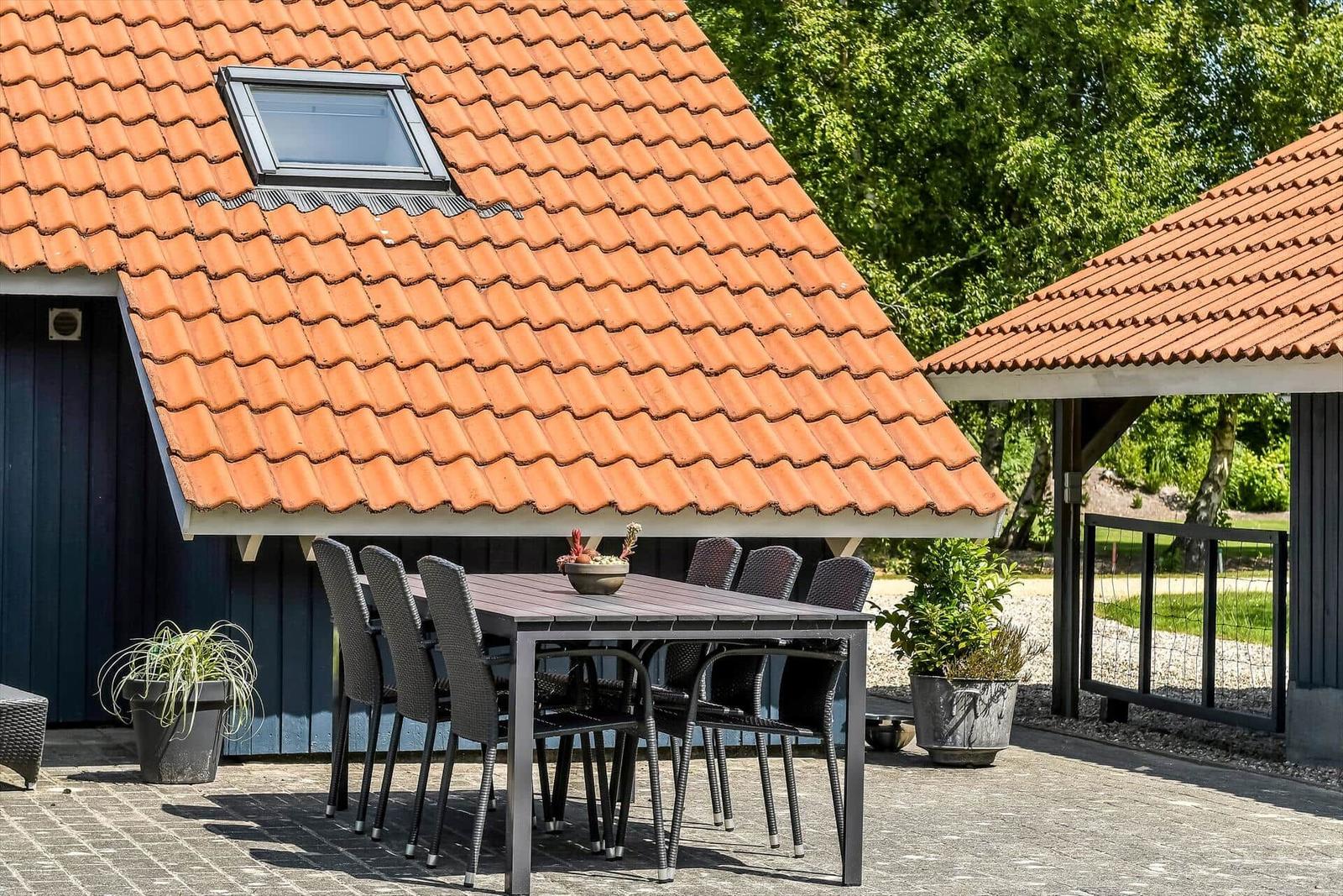 Terrasse mit Tisch und Stühlen unter rotem Ziegeldach. Pflanzen und Garten im Hintergrund.