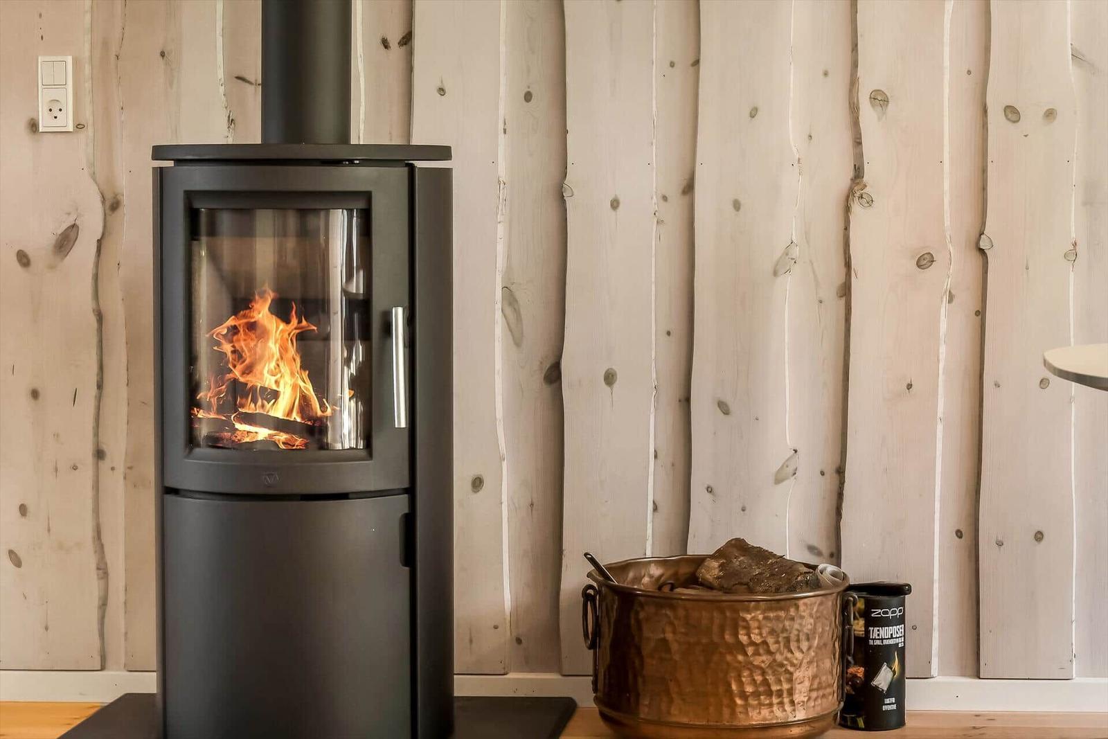 Holzofen mit brennendem Feuer an Holzwand. Kupferkessel und Brennstoffbehälter daneben.