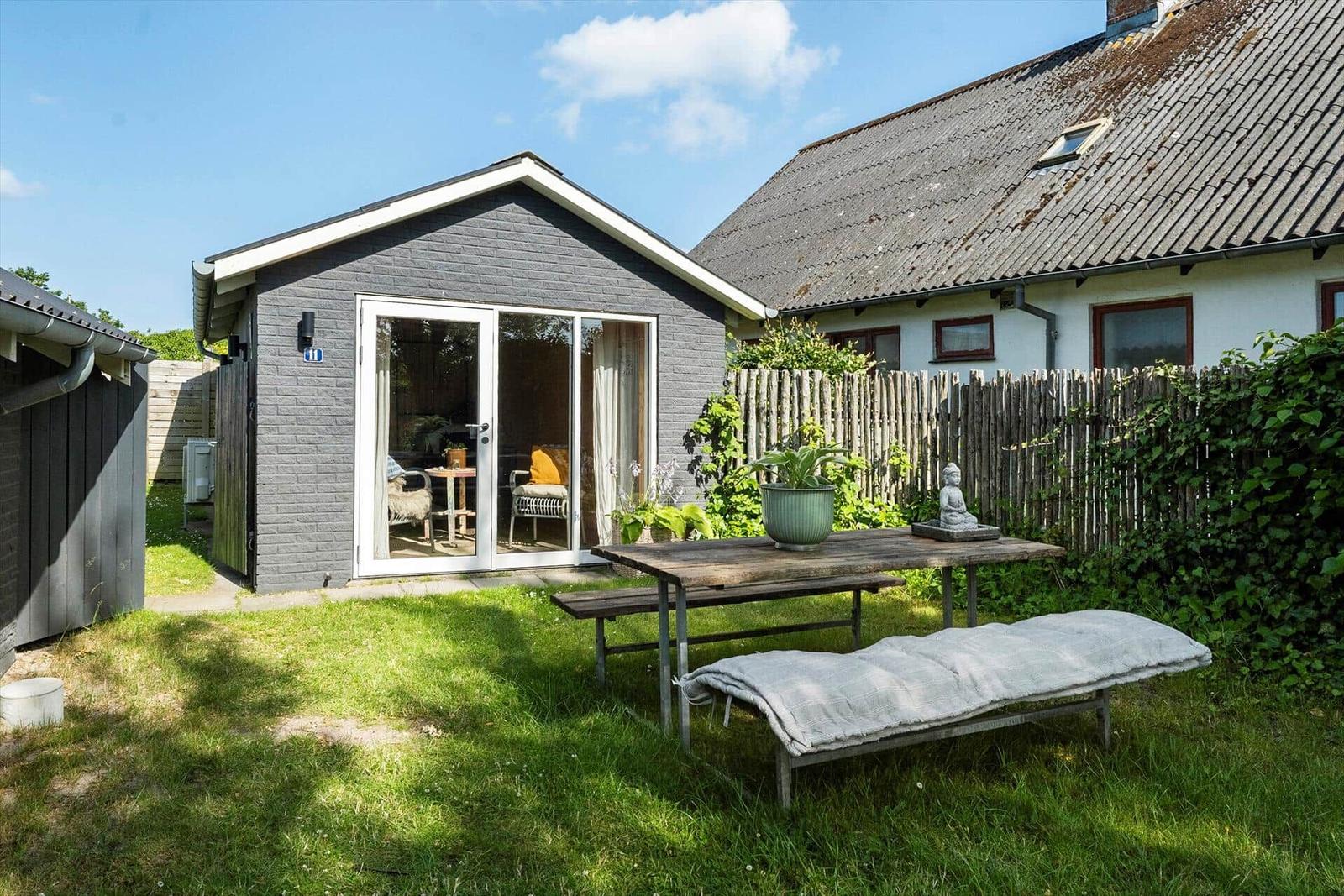 Garten mit Holztisch, Bank und Liege vor grauem Haus mit Glasfront.