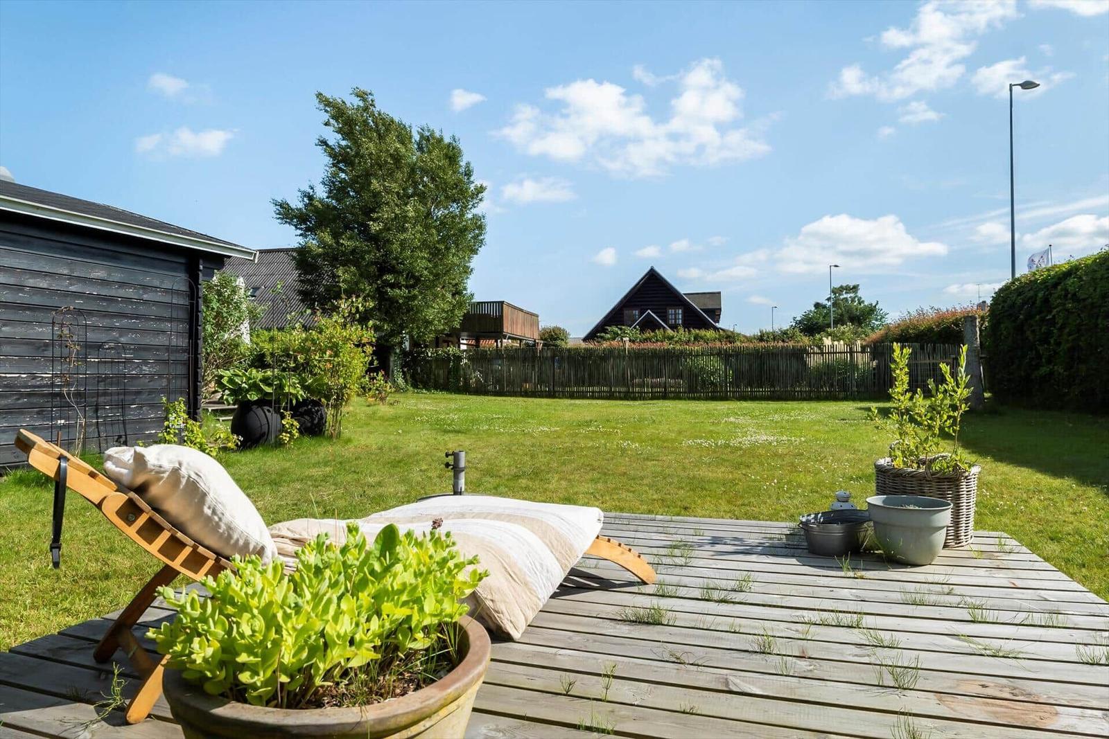 Hinterhof mit Holzterrasse, Liegestuhl und Grünflächen. Im Hintergrund sind Häuser und Bäume sichtbar.
