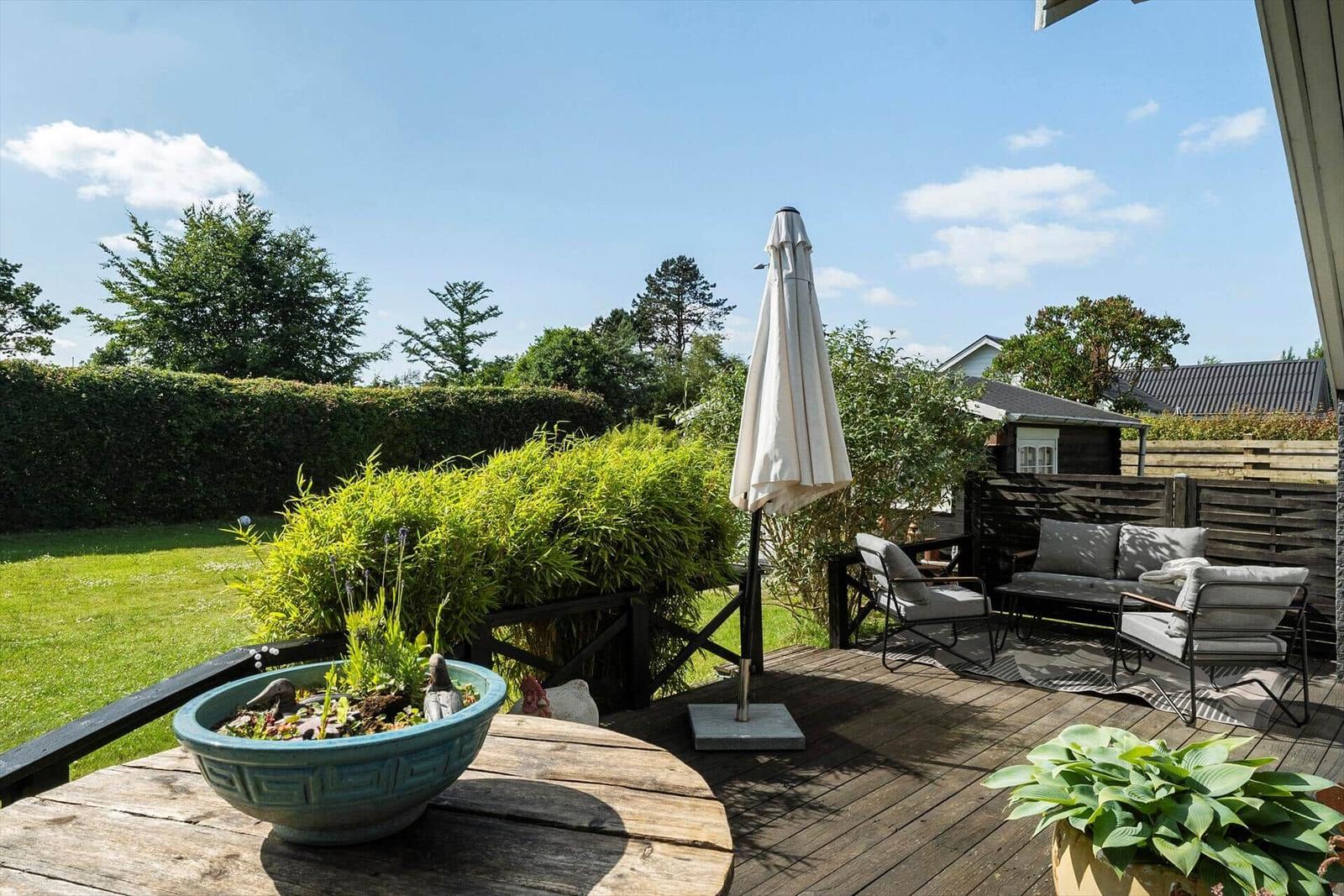 Terrasse mit Sitzgruppe, Sonnenschirm und Pflanzen. Grünfläche und Bäume im Hintergrund.