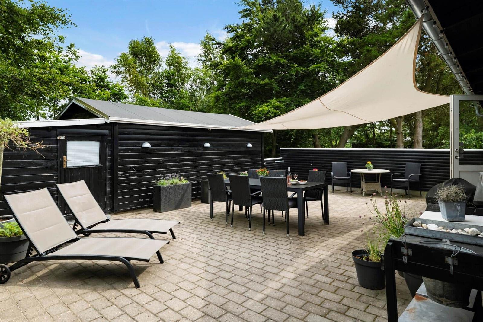 Terrasse mit Sonnensegel, Liegestühlen und Esstisch. Hintergrund: schwarzes Holzhaus und Bäume.