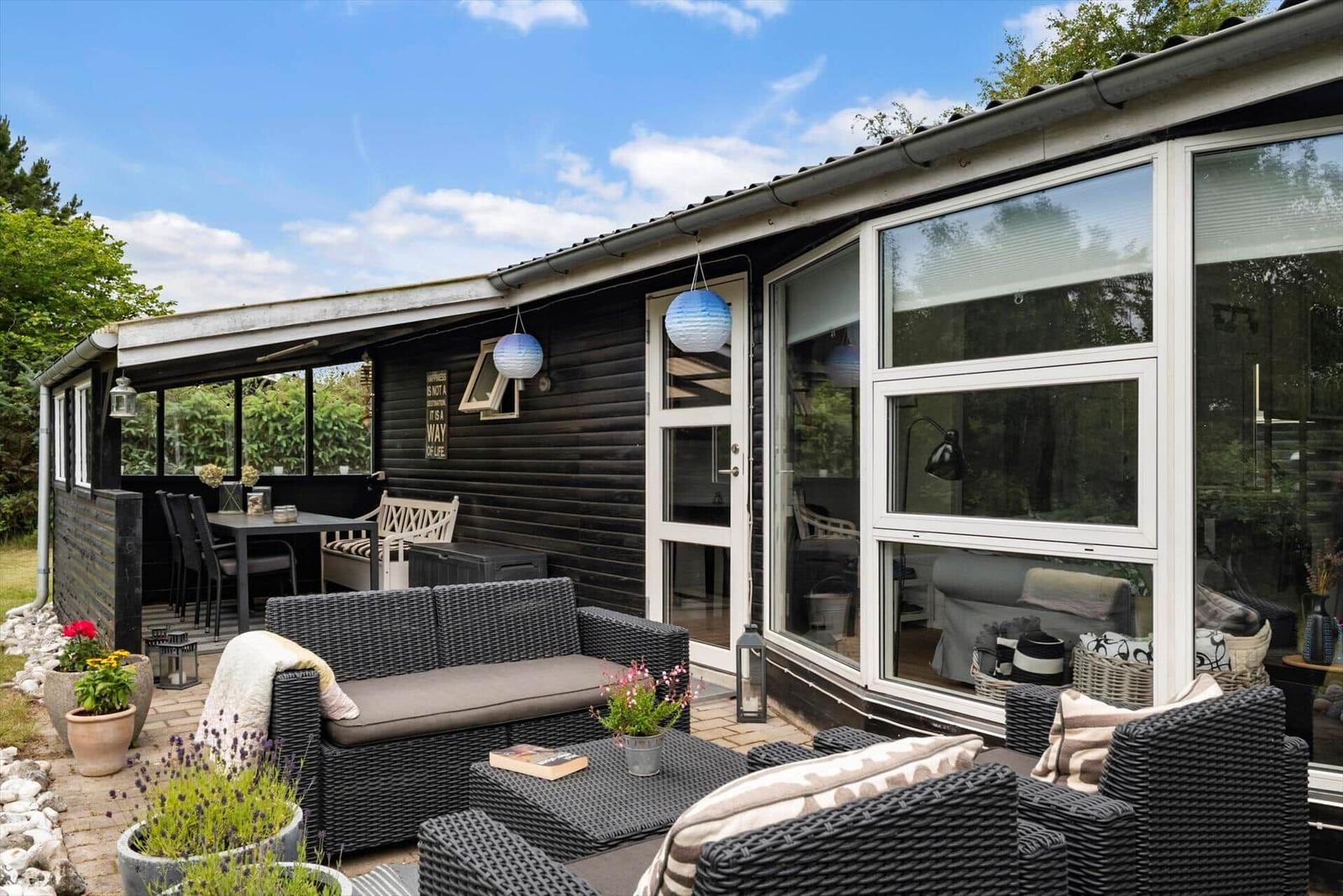 Terrasse mit Sitzgruppe, Tisch und Blumen. Haus mit Glasfront und Holzwand.