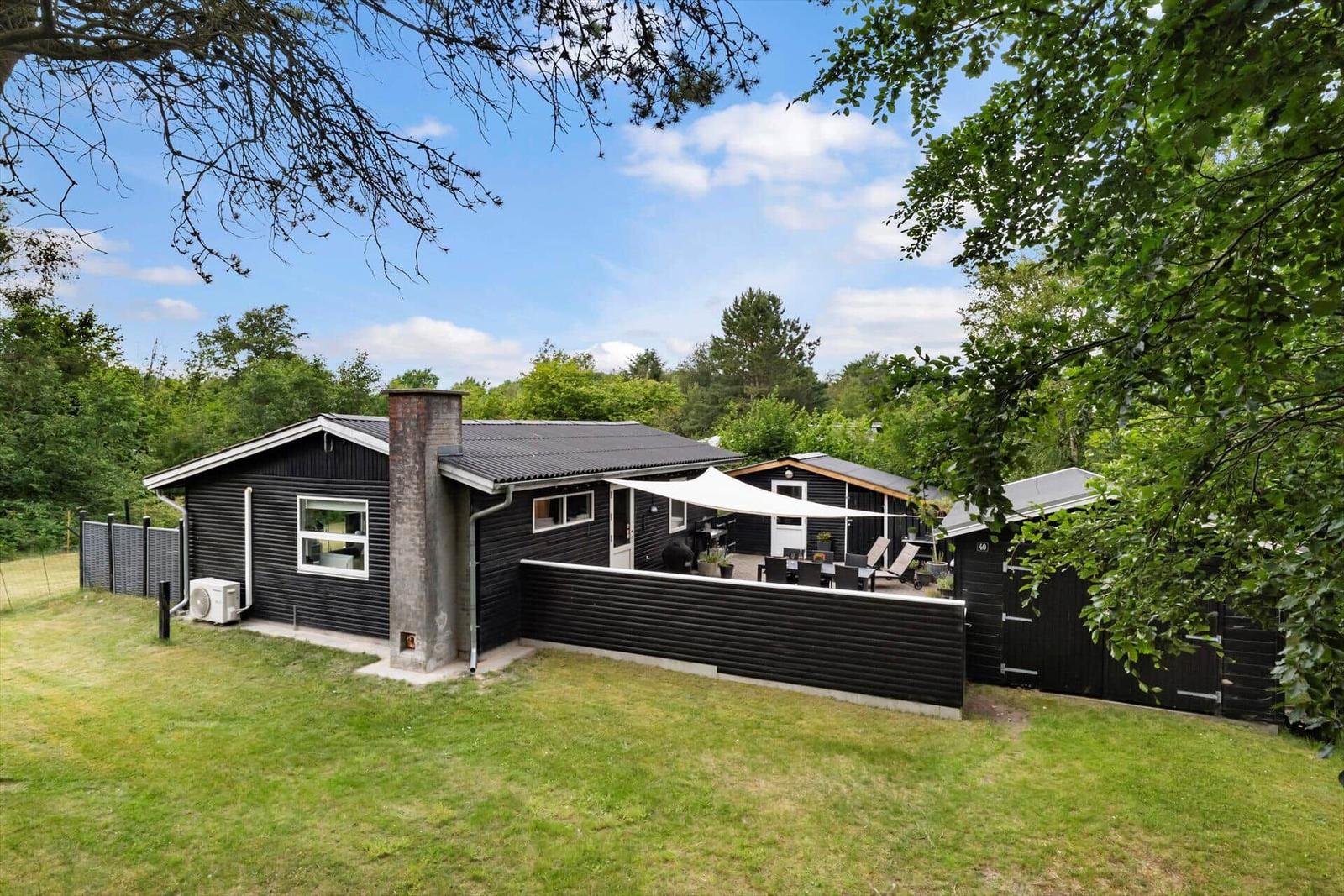 Schwarzes Ferienhaus mit Terrasse, Sonnenschutz und Garten. Umgeben von Bäumen und Grün.