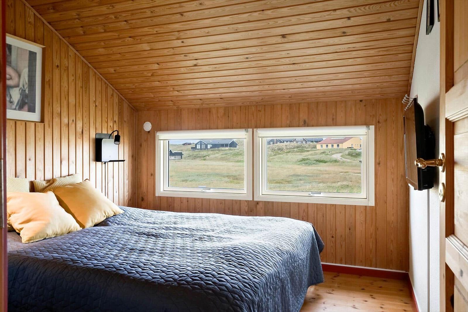 Schlafzimmer mit Holzwand und -decke, Bett mit blauer Bettwäsche und zwei Fenstern mit Blick auf Landschaft.