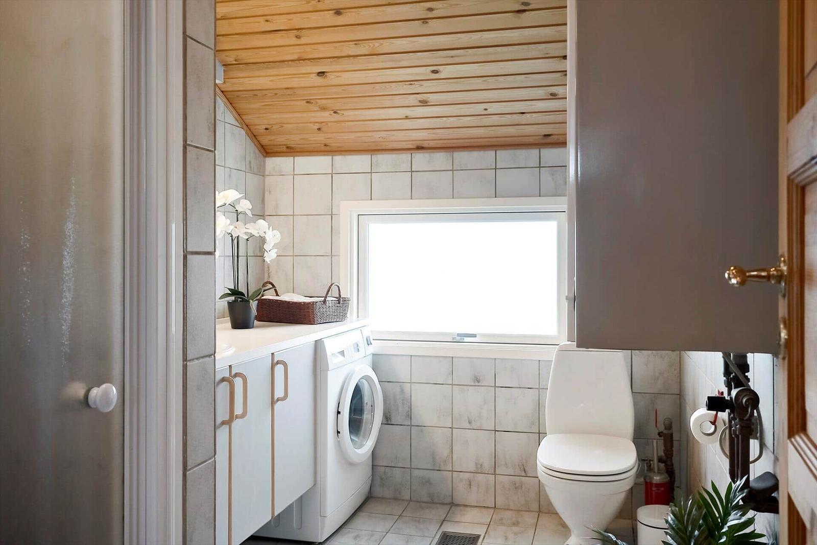 Badezimmer mit Waschmaschine, Toilette und Fenster. Holzdecke und Fliesenwand.