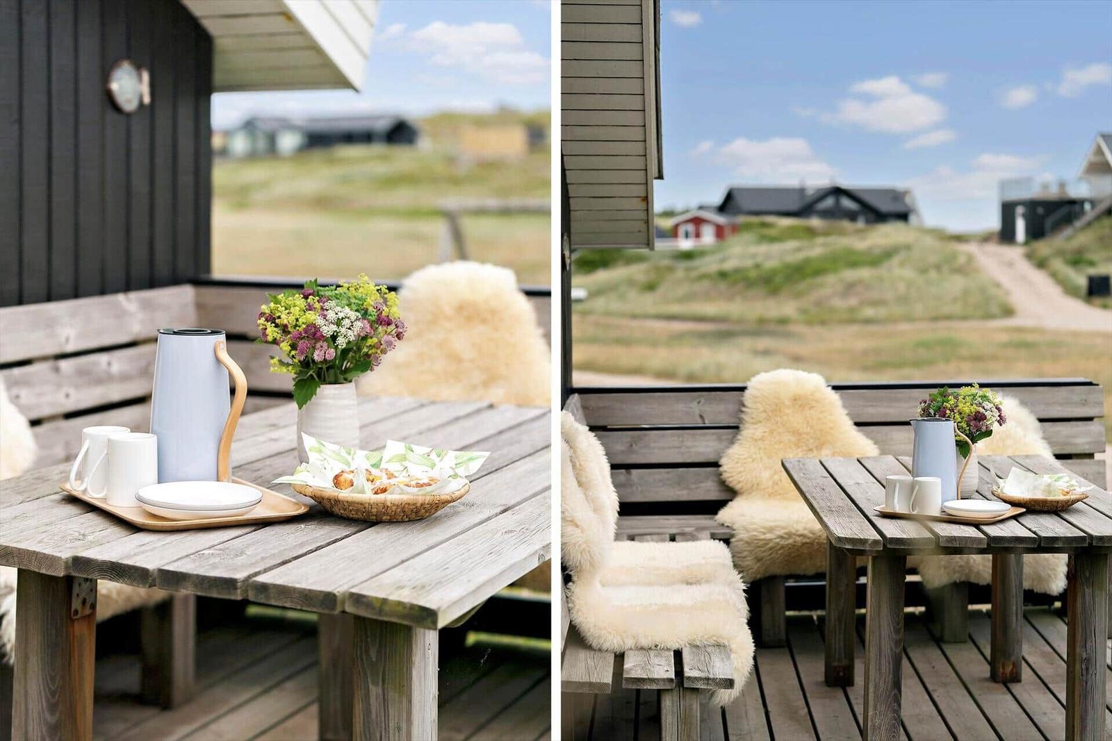 Terrasse mit Holztisch, Stühlen und Frühstückszubehör. Blick auf grüne Dünen.