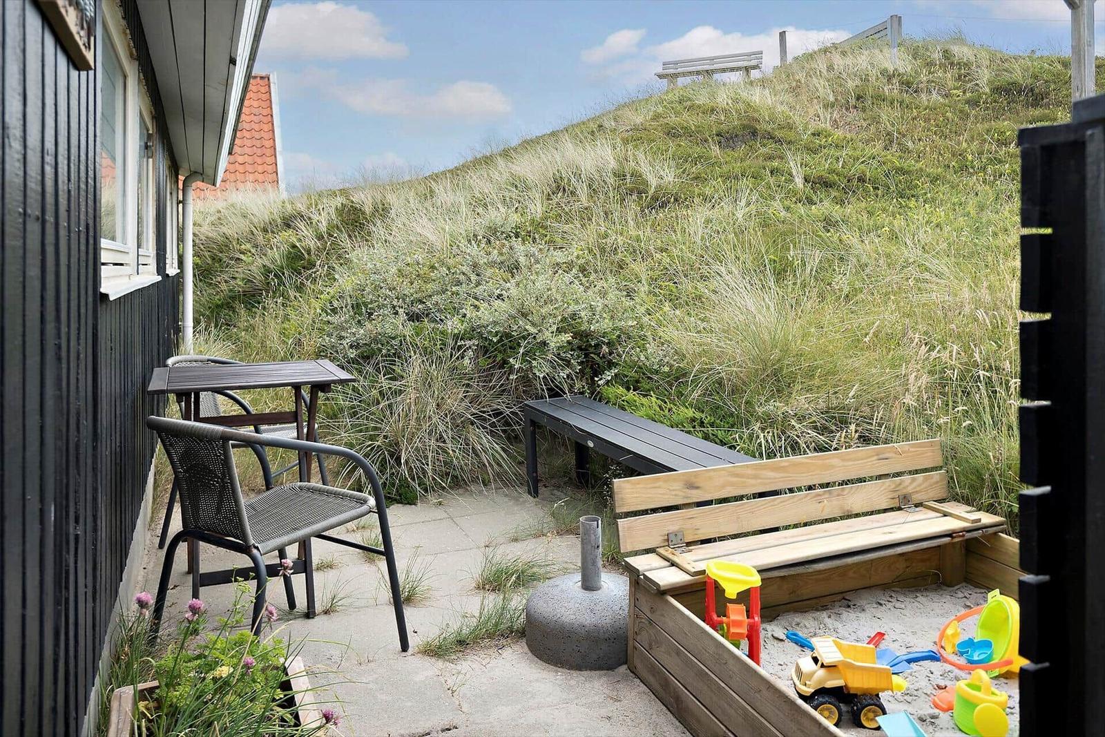 Terrasse mit Tisch, Stühlen, Sandkasten und Blick auf Grashügel.