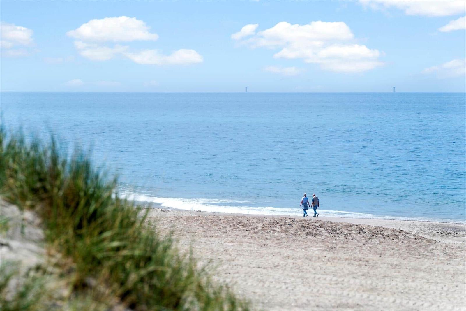 Zwei Personen spazieren am Strand entlang, im Hintergrund das Meer und ein Windrad.