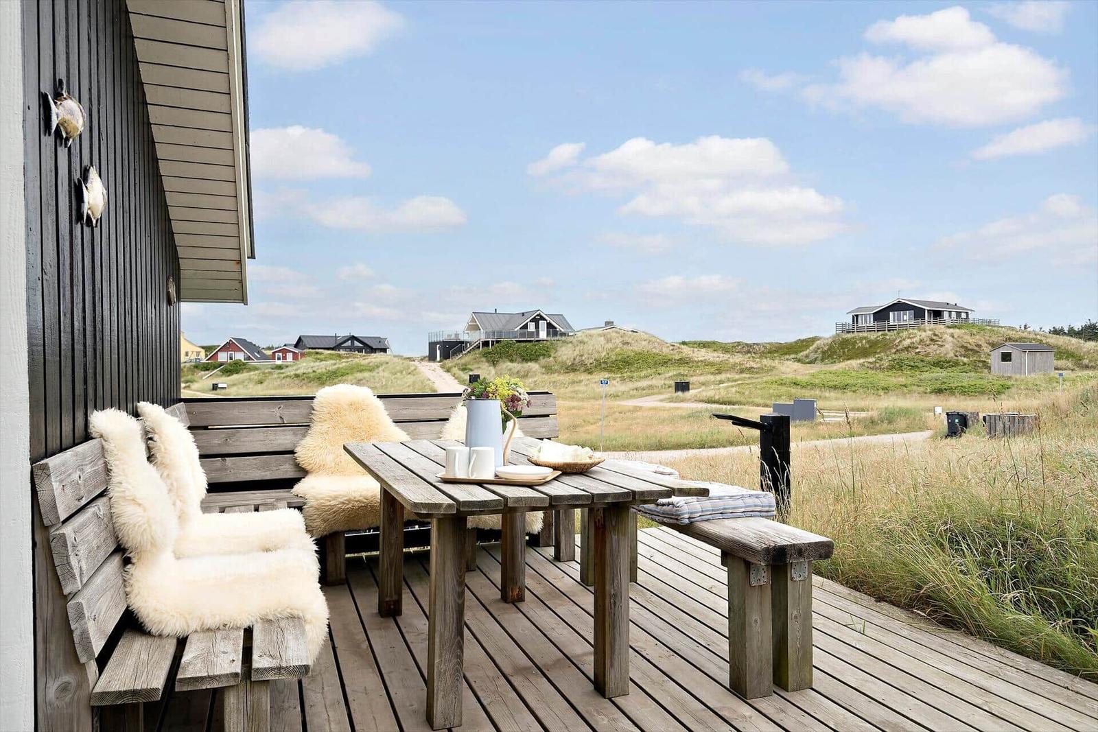 Terrasse mit Holztisch und Bänken, weiß gefüttert, Blick auf Dünen und Häuser.