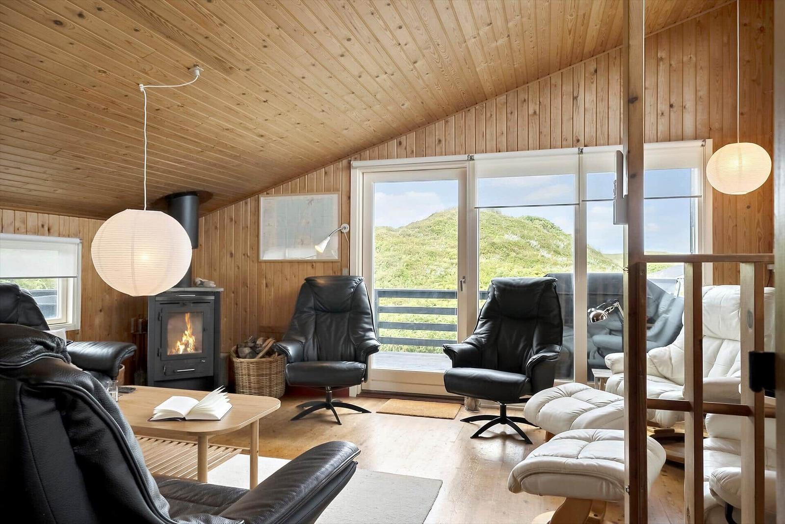 Wohnzimmer mit Holzwand, Kamin und Sitzgelegenheiten. Blick auf grüne Hügel.