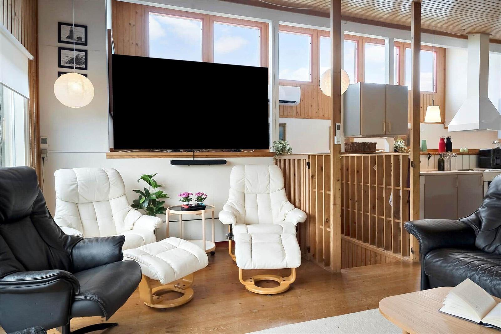 Wohnzimmer mit Fernseher, weißen Sesseln und Holzelementen. Küche im Hintergrund.