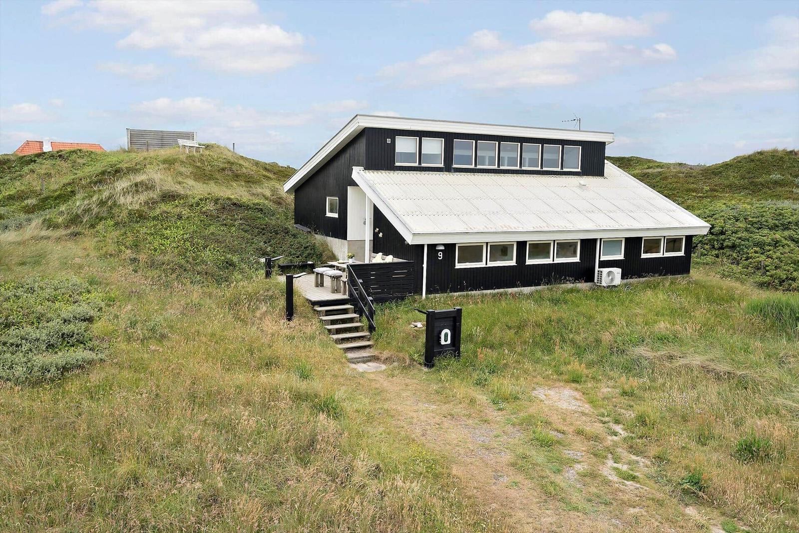 Schwarzes Haus mit weißem Dach auf grüner Dünenlandschaft. Außenbereich mit Tisch und Stufen.