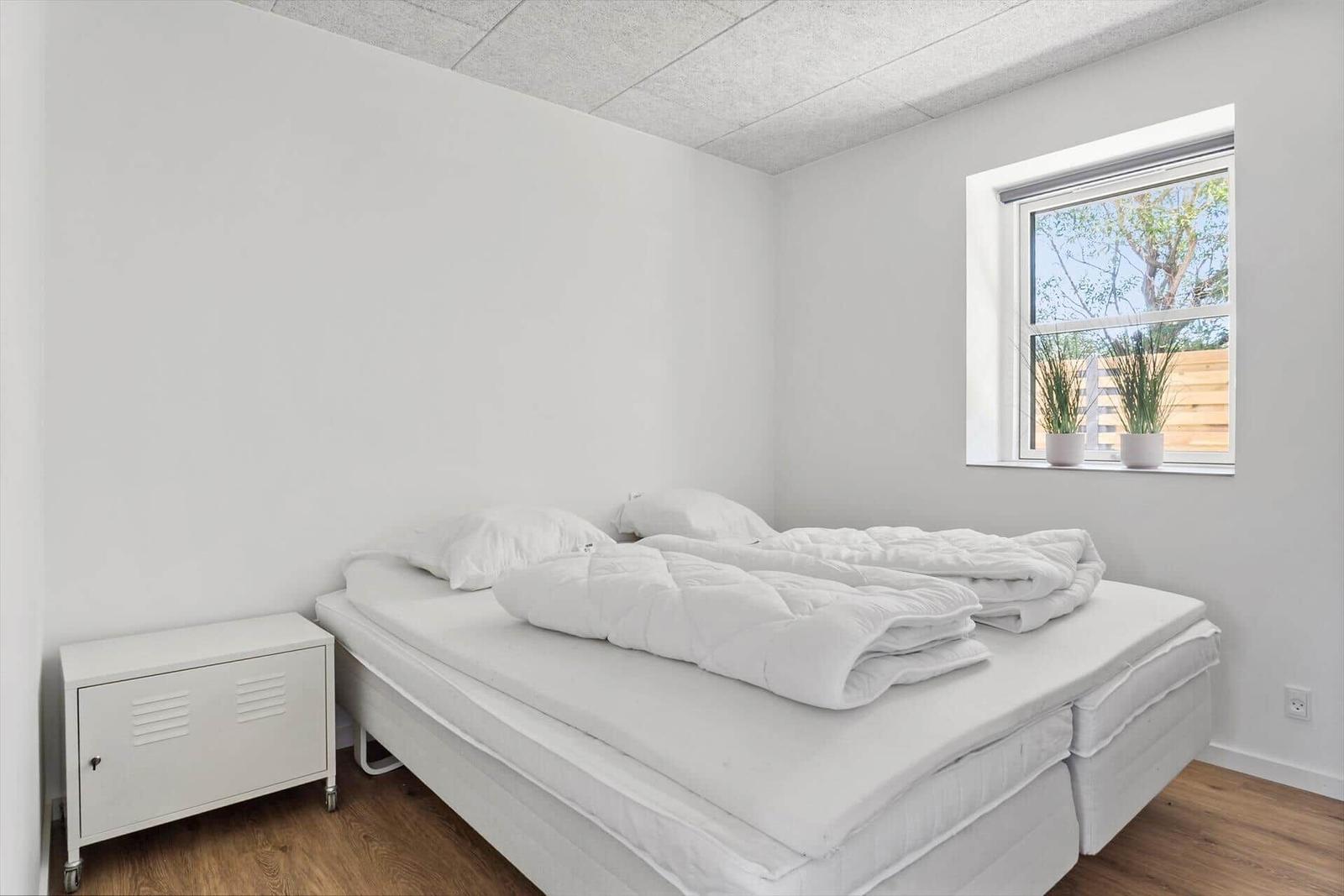 Schlafzimmer mit weißem Bett, Nachttisch und Fenster mit Pflanzen.