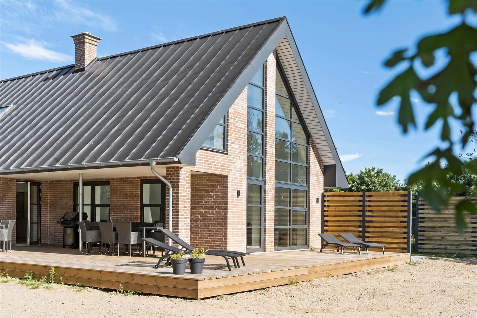 Moderne Villa mit Holzterrasse, Grill und Liegestühlen. Große Fensterfront und Ziegelmauer.