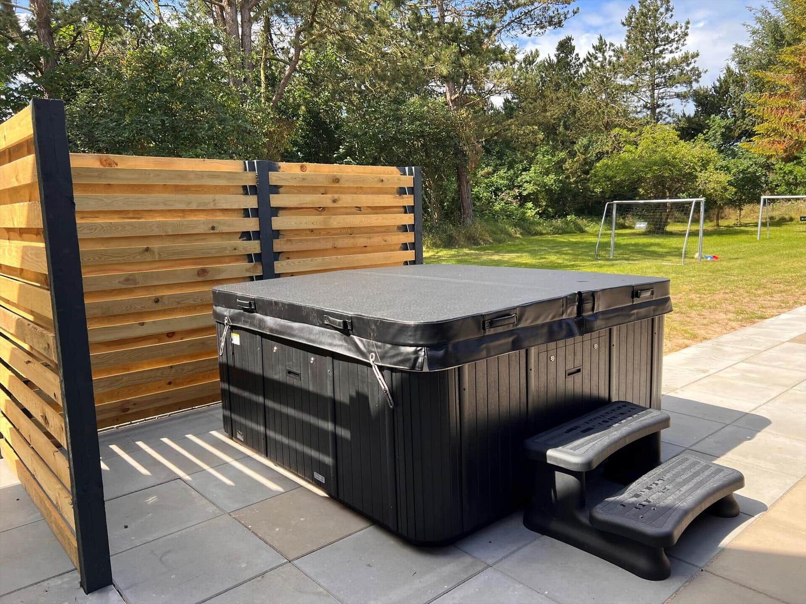 Schwarzes Whirlpoolbecken auf Terrasse mit Holzumrandung und Grünfläche im Hintergrund.
