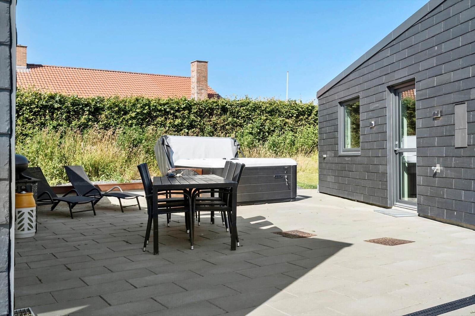 Terrasse mit Tisch, Stühlen, Liegestühlen und Whirlpool. Hintergrund: grüner Heckenzaun und Haus.