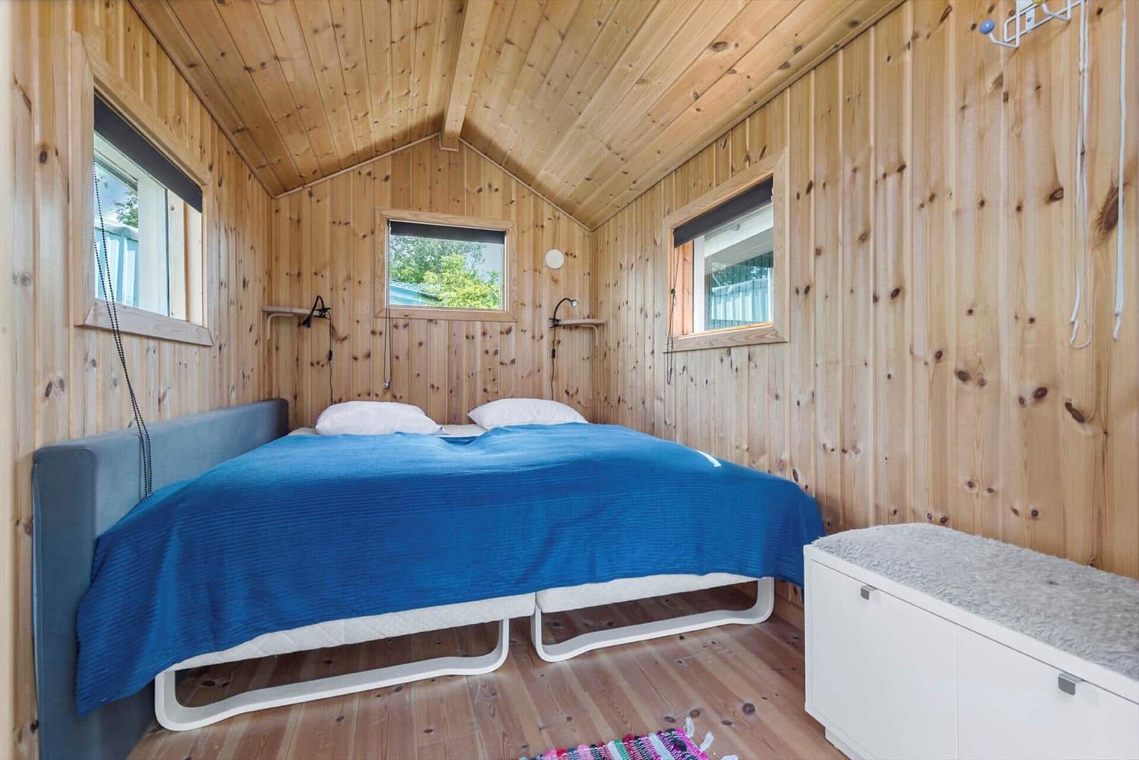 Schlafzimmer mit Holzwand, Bett mit blauer Bettdecke und drei Fenstern.