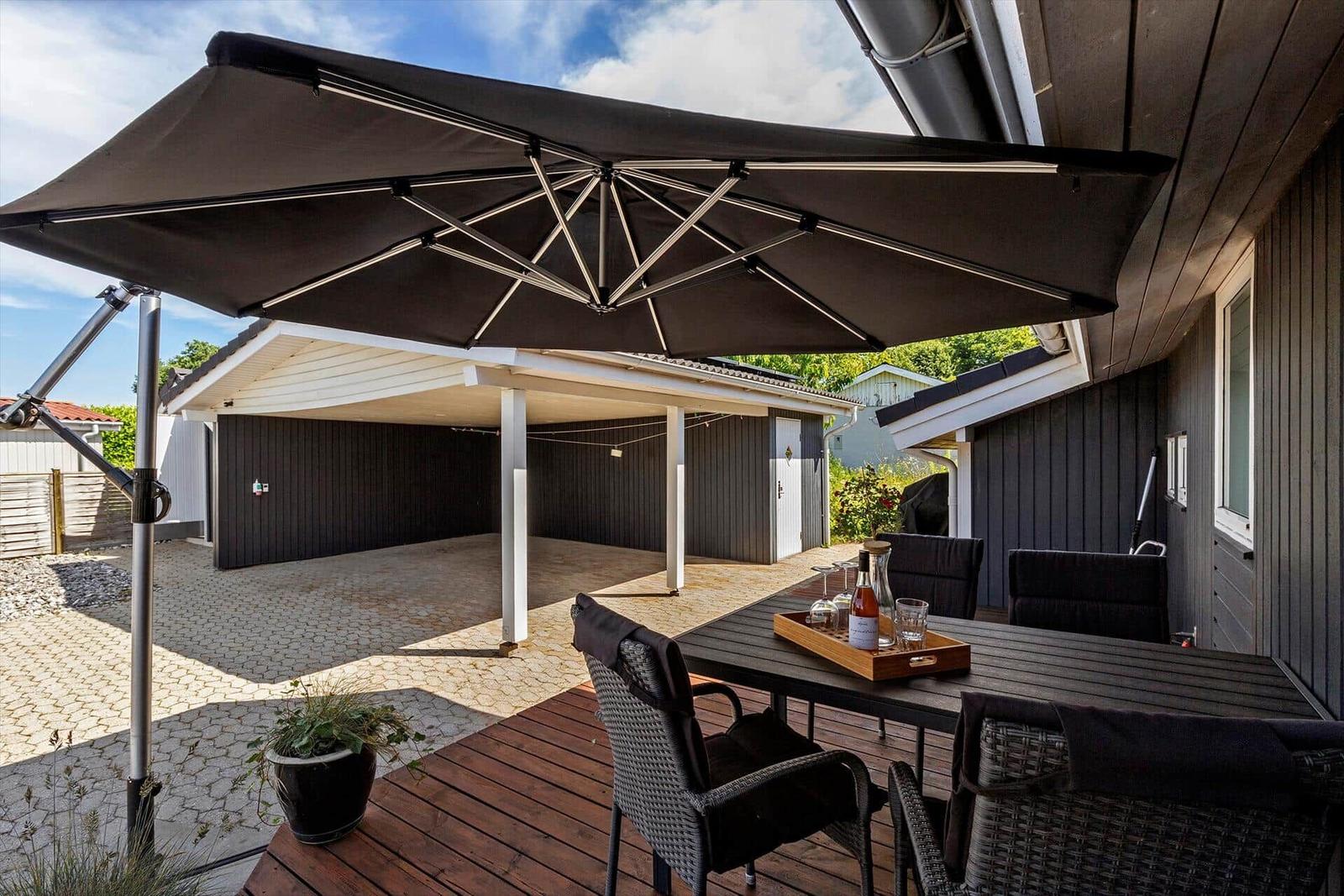 Terrasse mit Tisch, Stühlen und Sonnenschutz. Hintergrund: Carport und Holzhaus.