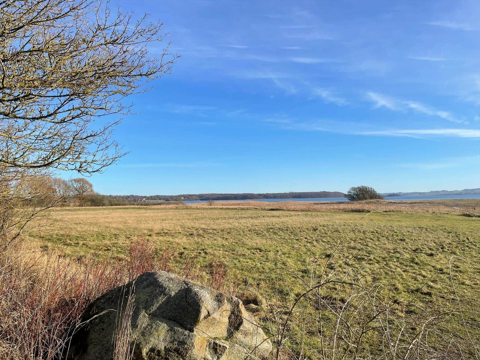Weitläufiges Grasland mit Stein und Blick auf einen See unter blauem Himmel.
