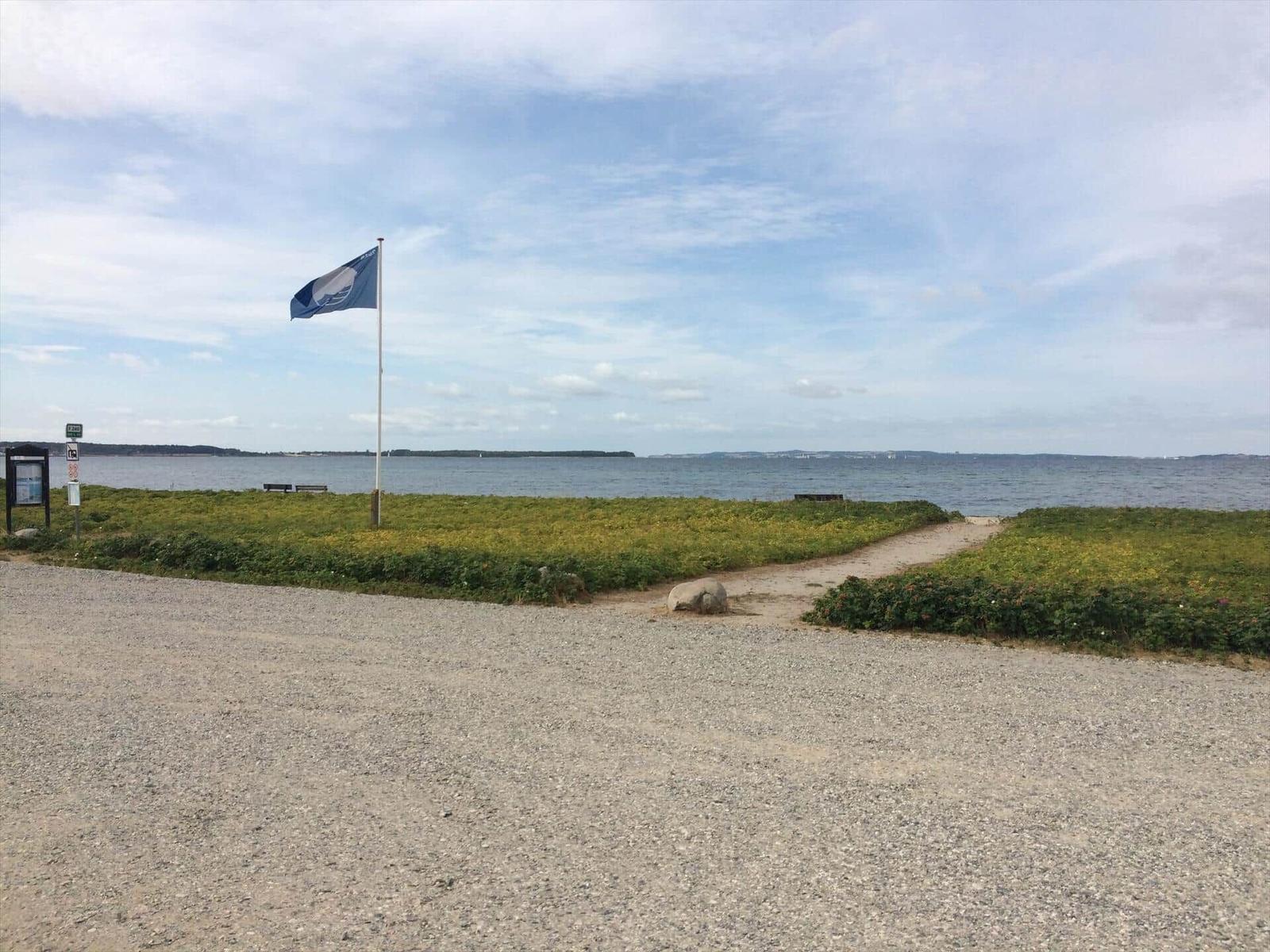 Grauer Kiesweg führt zu einem Uferbereich mit Flagge und Grünflächen.