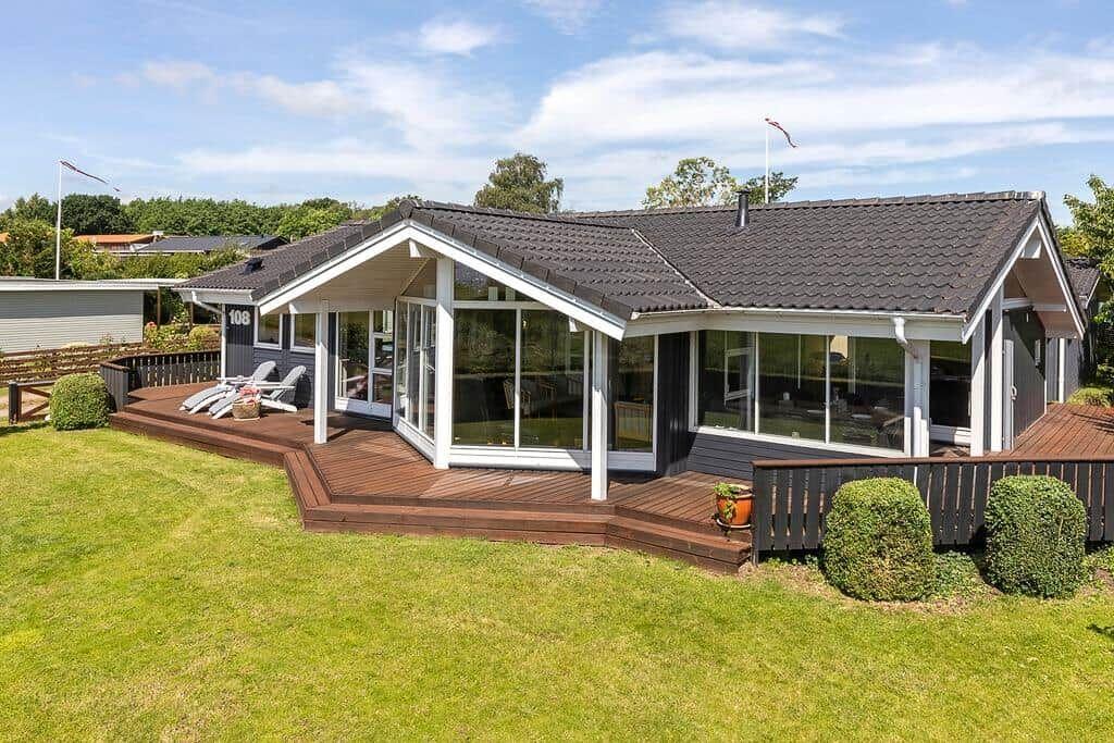 Moderne Villa mit großem Holzterrasse und Glasfront. Grünfläche und Bäume umgeben das Haus.