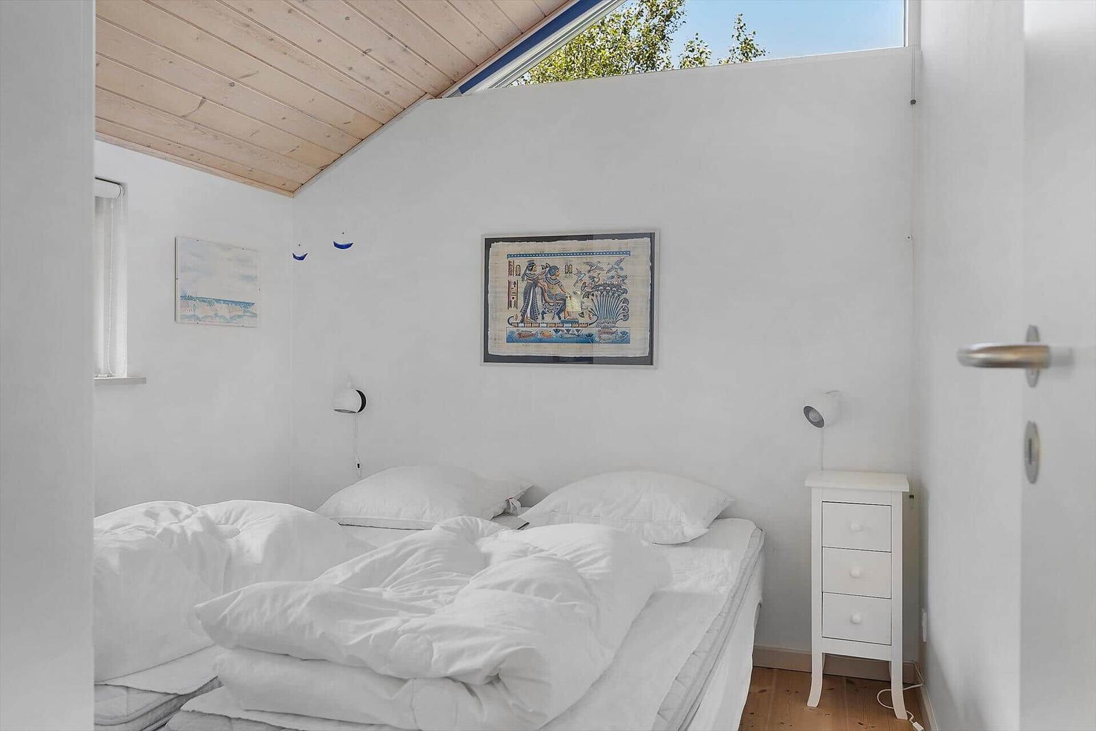 Schlafzimmer mit weißem Bett, Nachttisch und Wandbild. Holzdecke und Fenster.