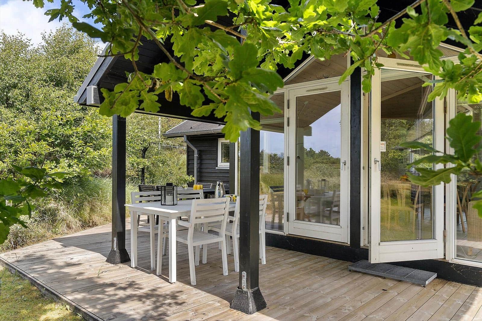 Terrasse mit weißem Tisch und Stühlen unter schwarzem Dach. Glasfront öffnet sich zum Innenraum.