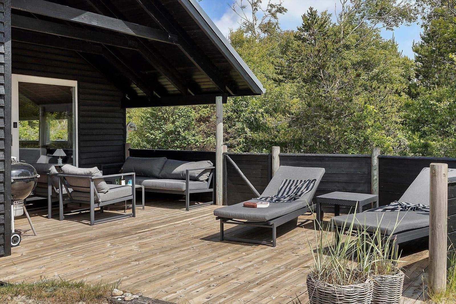 Terrasse mit Liegen, Sofa und Grill unter schwarzem Dach. Hintergrund: grüne Bäume.