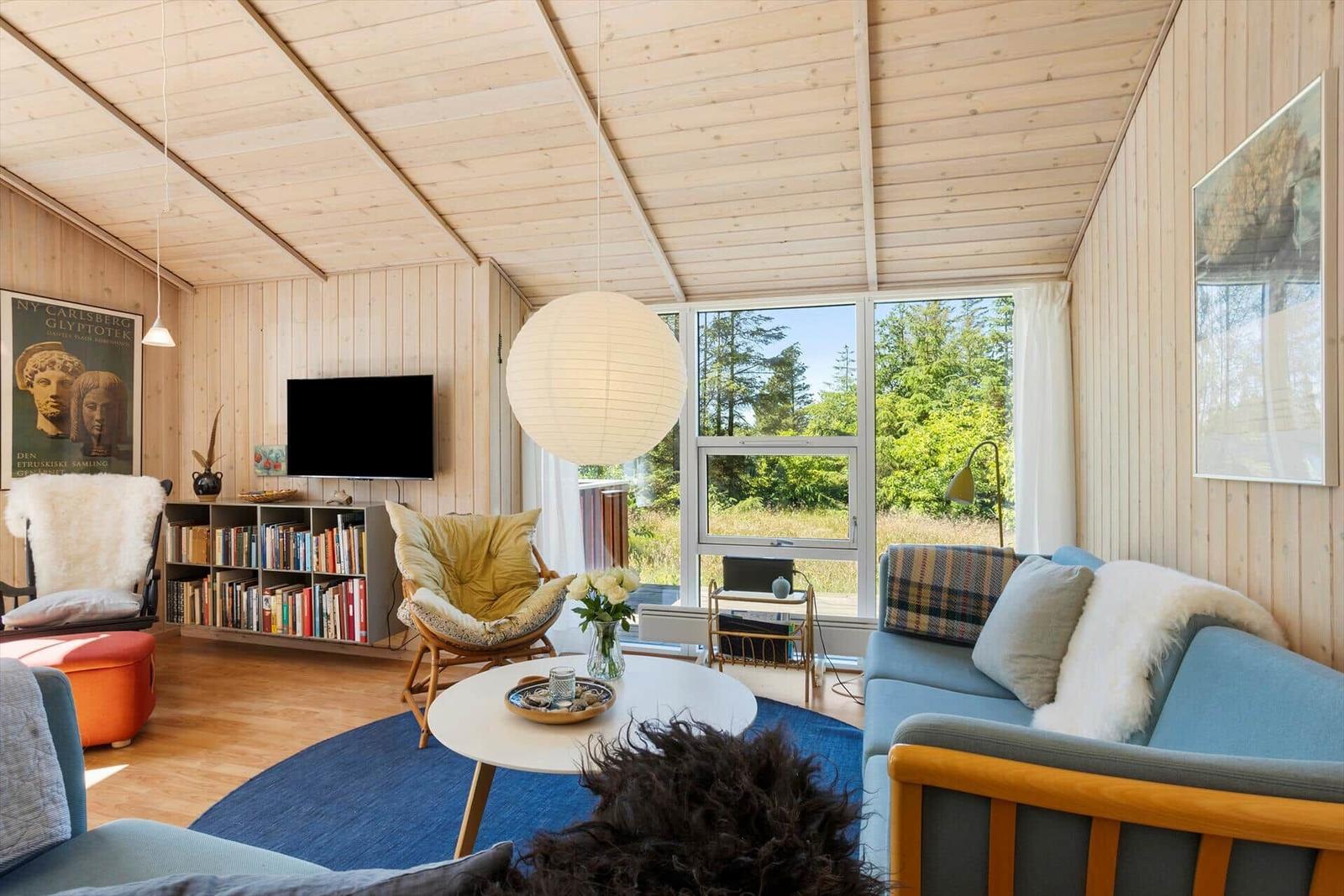 Living room with wood walls, sofa, chairs, and large window overlooking forest.