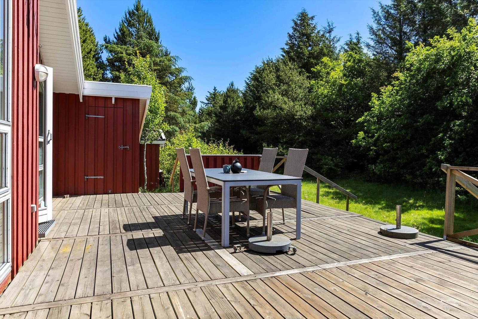 Deck with table and chairs beside red house with view of green trees.