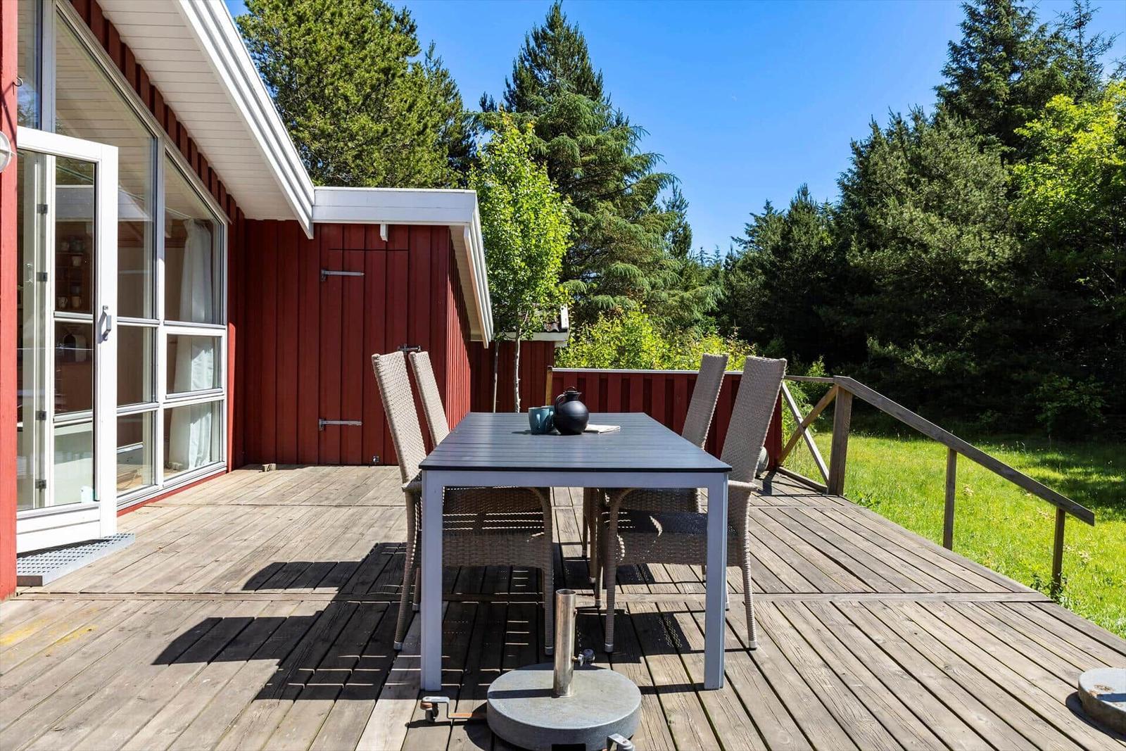 Deck with table and chairs in front of red house. Surrounded by green with wooden railing.