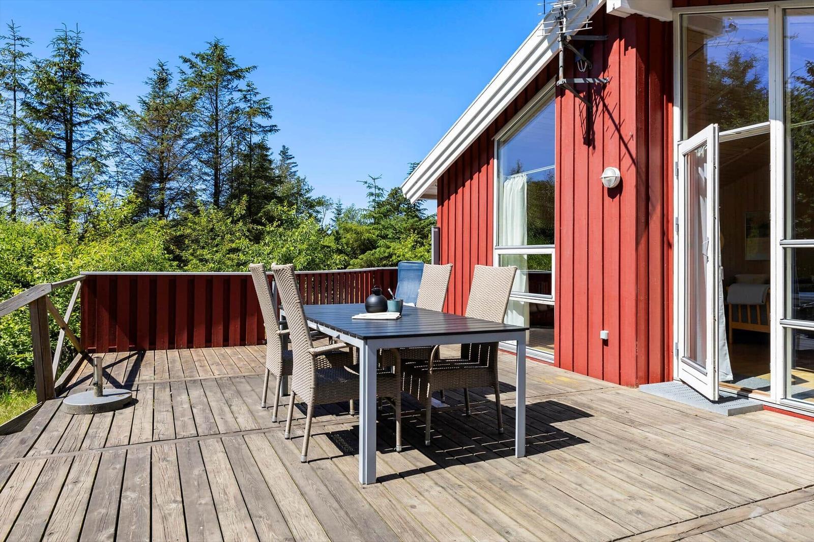 Deck with table and chairs in front of red house with forest view.
