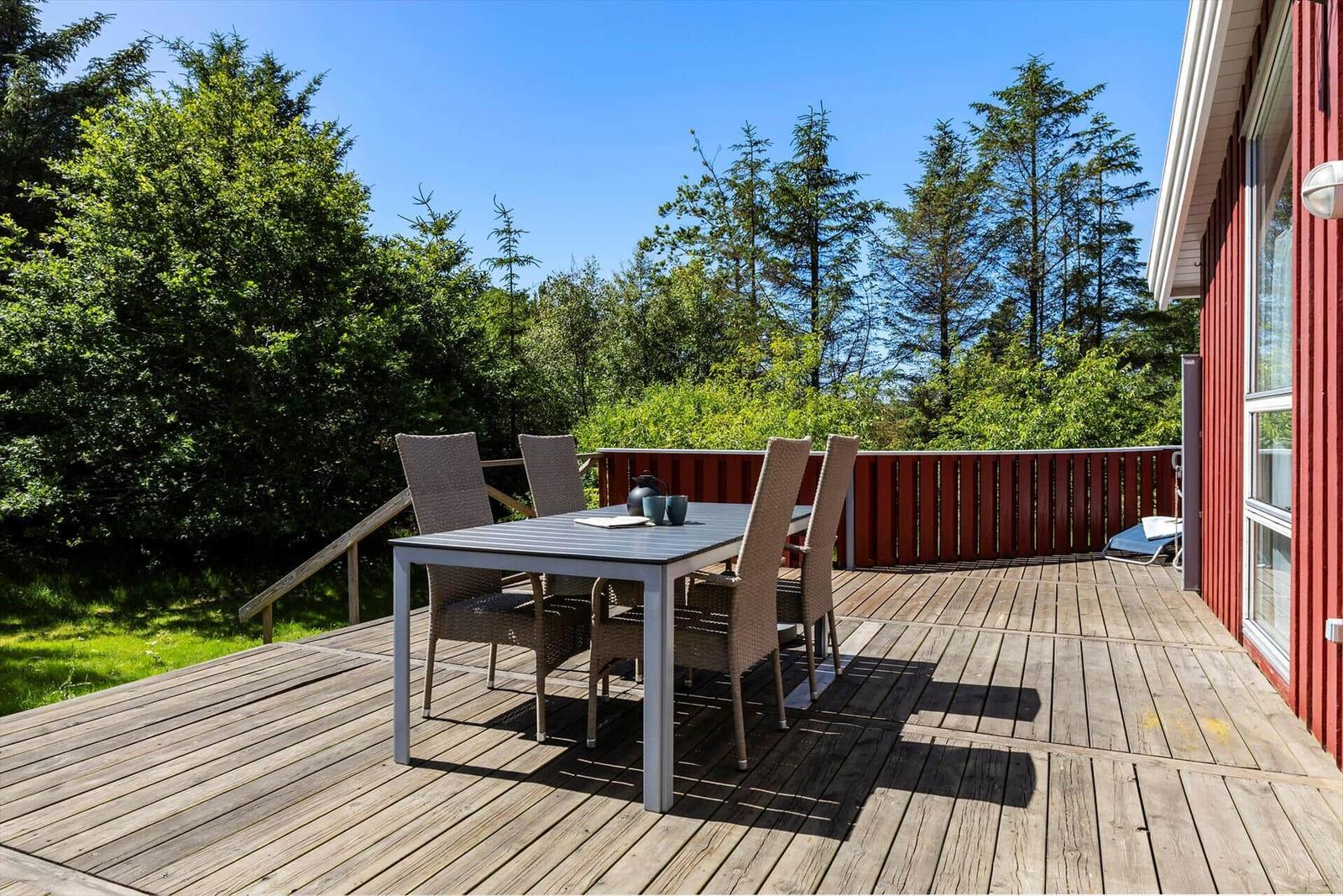 Deck with table and chairs next to red house. View into green trees.