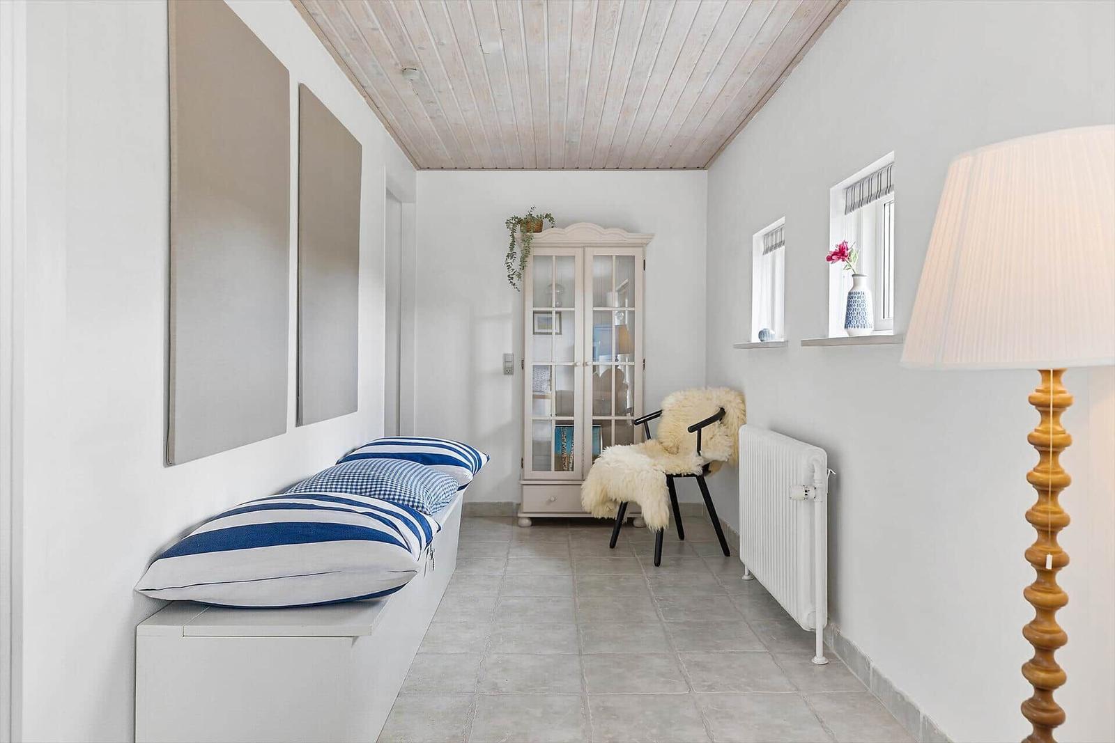 Langgezogener Raum mit Sitzbank, Schrank und Stehlampe. Holzdecke und Fenster mit Blumen.