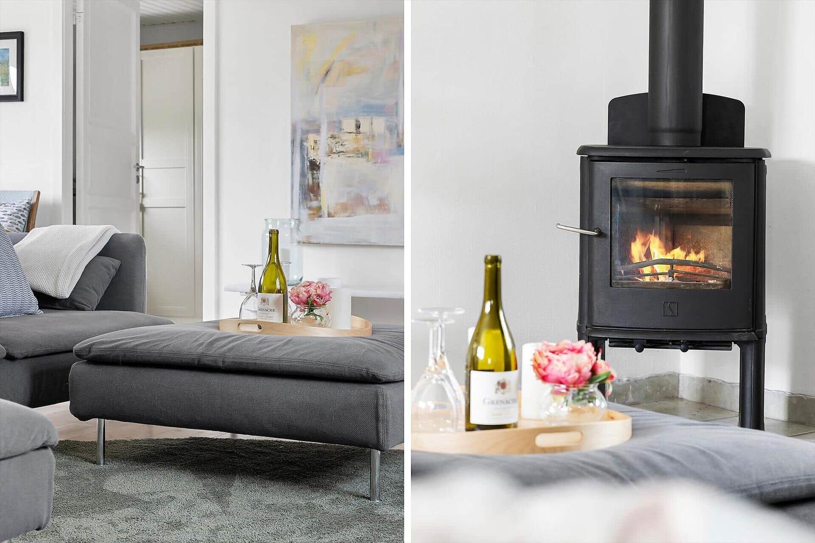 Ein Wohnzimmer mit grauem Sofa und Kaminofen. Auf einem Tisch stehen Wein und Blumen.