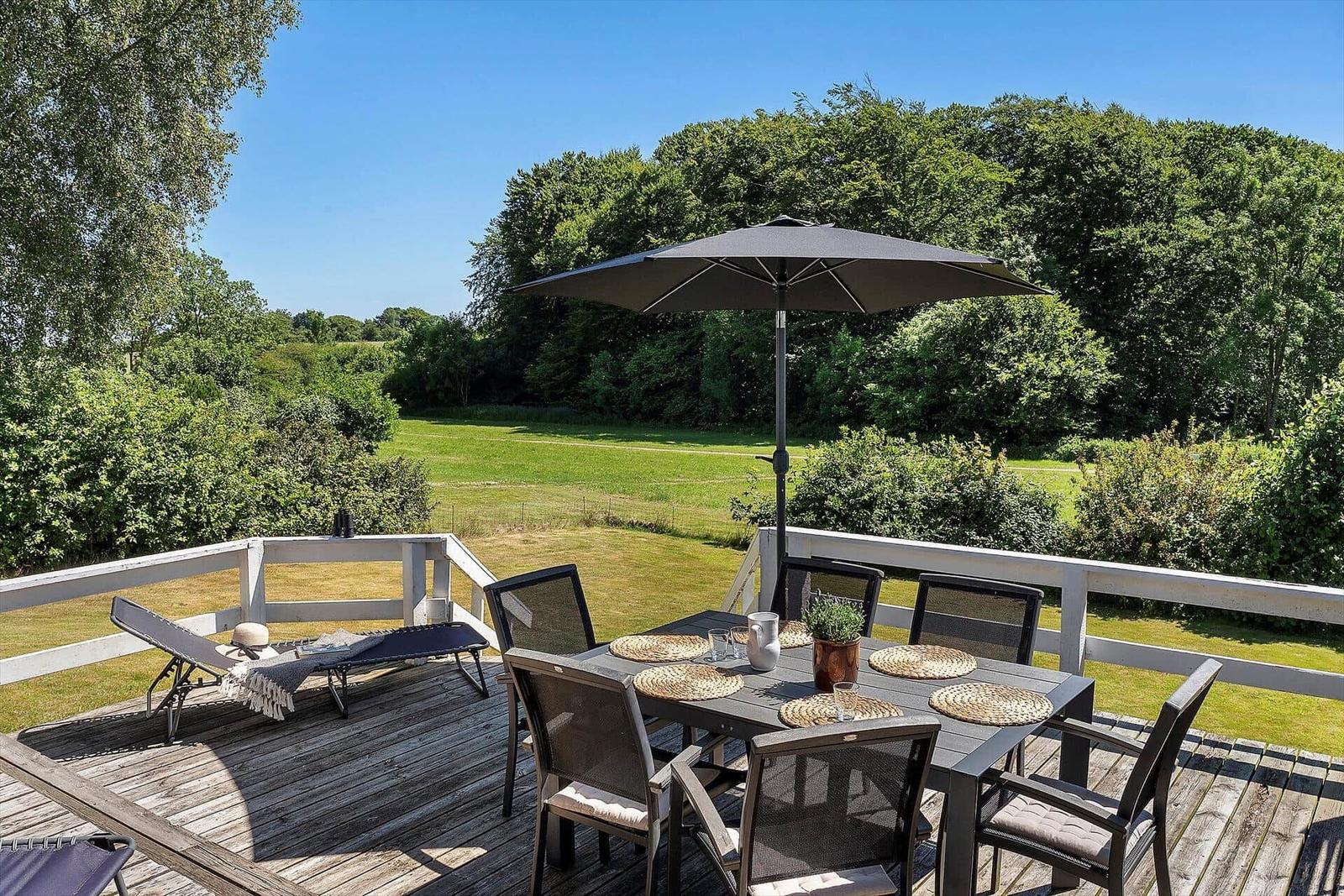 Deck with table, chairs, and umbrella overlooking green field.
