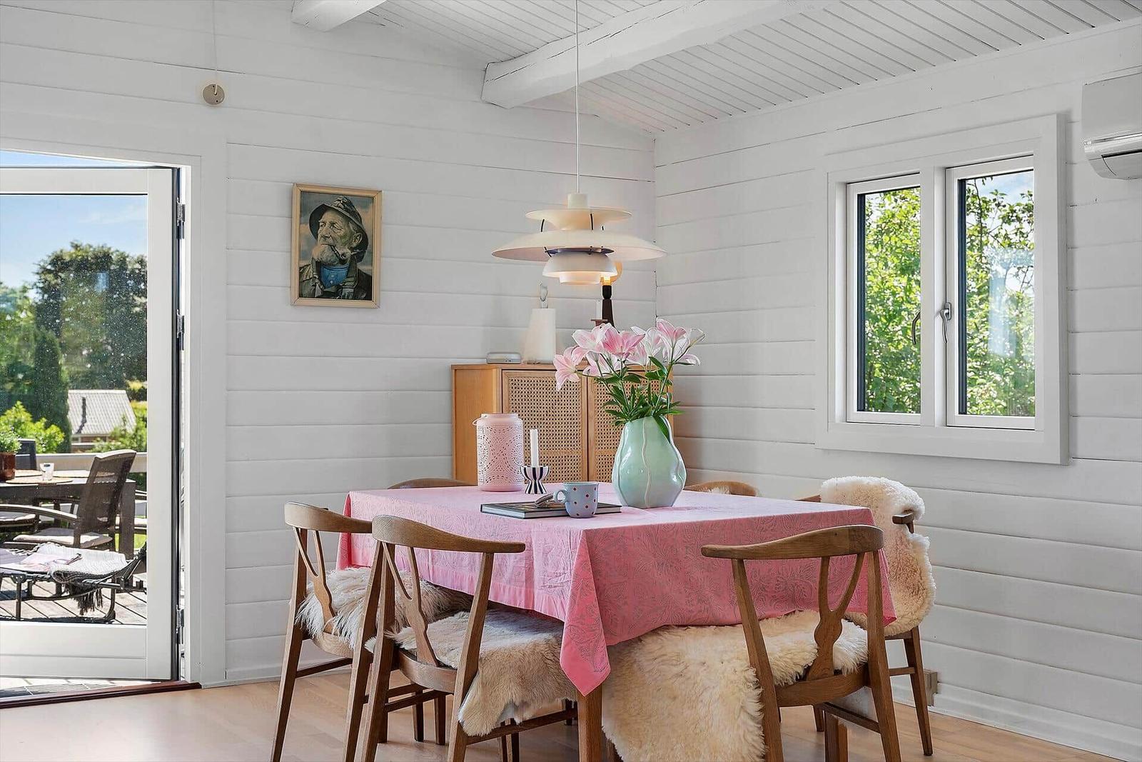 Dining room with wooden table, chairs, and pink tablecloth. White walls and windows with outdoor view.
