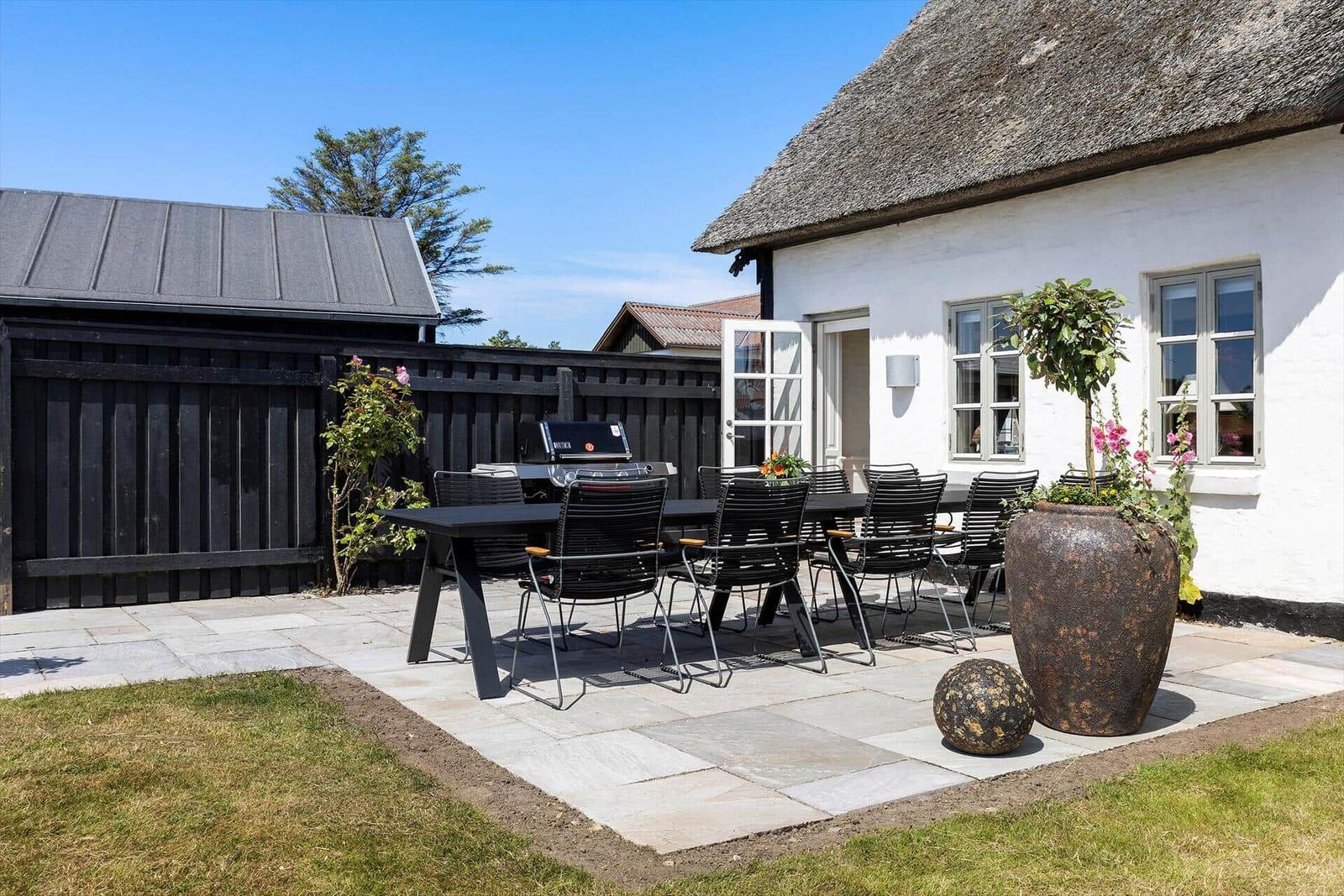 Terrace with table, chairs, and grill beside white house with thatched roof.