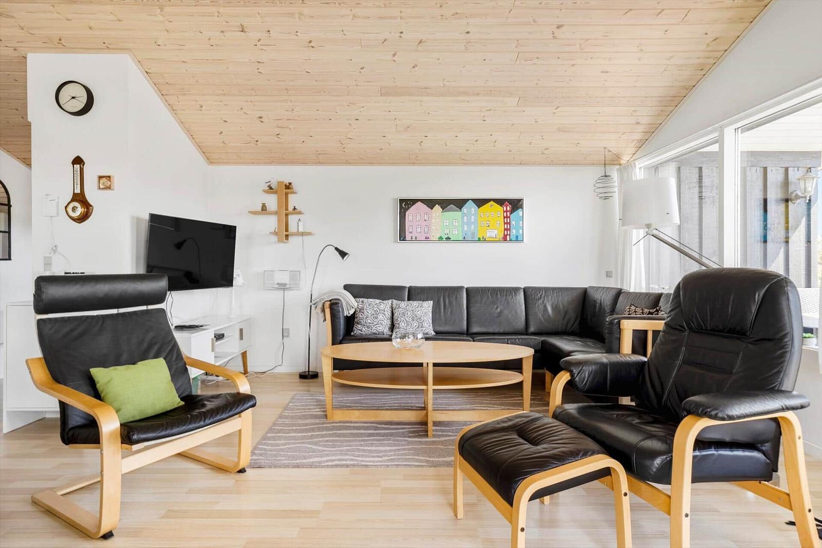 Wohnzimmer mit schwarzer Ledercouch, zwei Sessel und Holztisch. Holzdecke und Fensterfront.