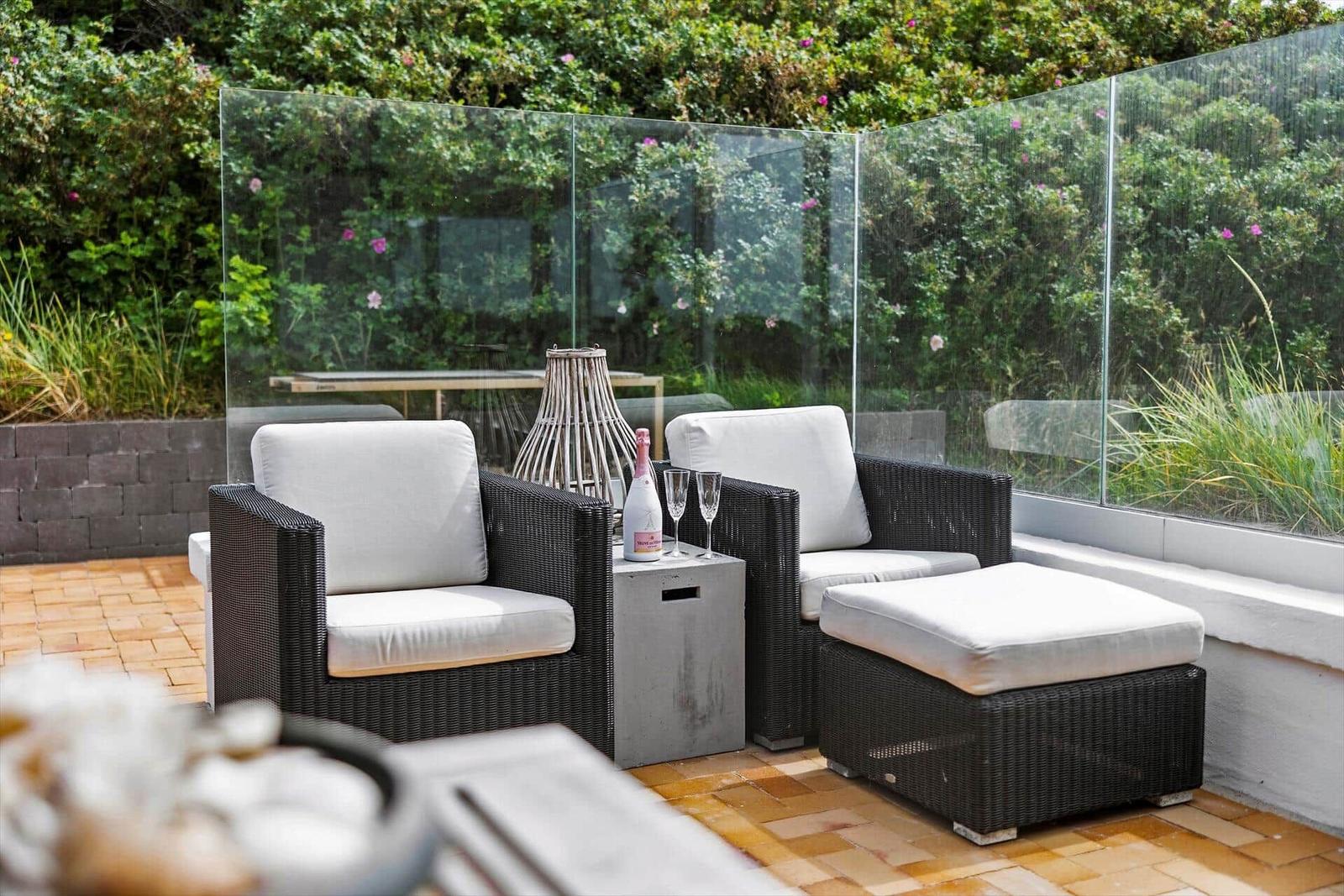 Terrace with two chairs, ottoman, and table. Glass wall and plants in background.